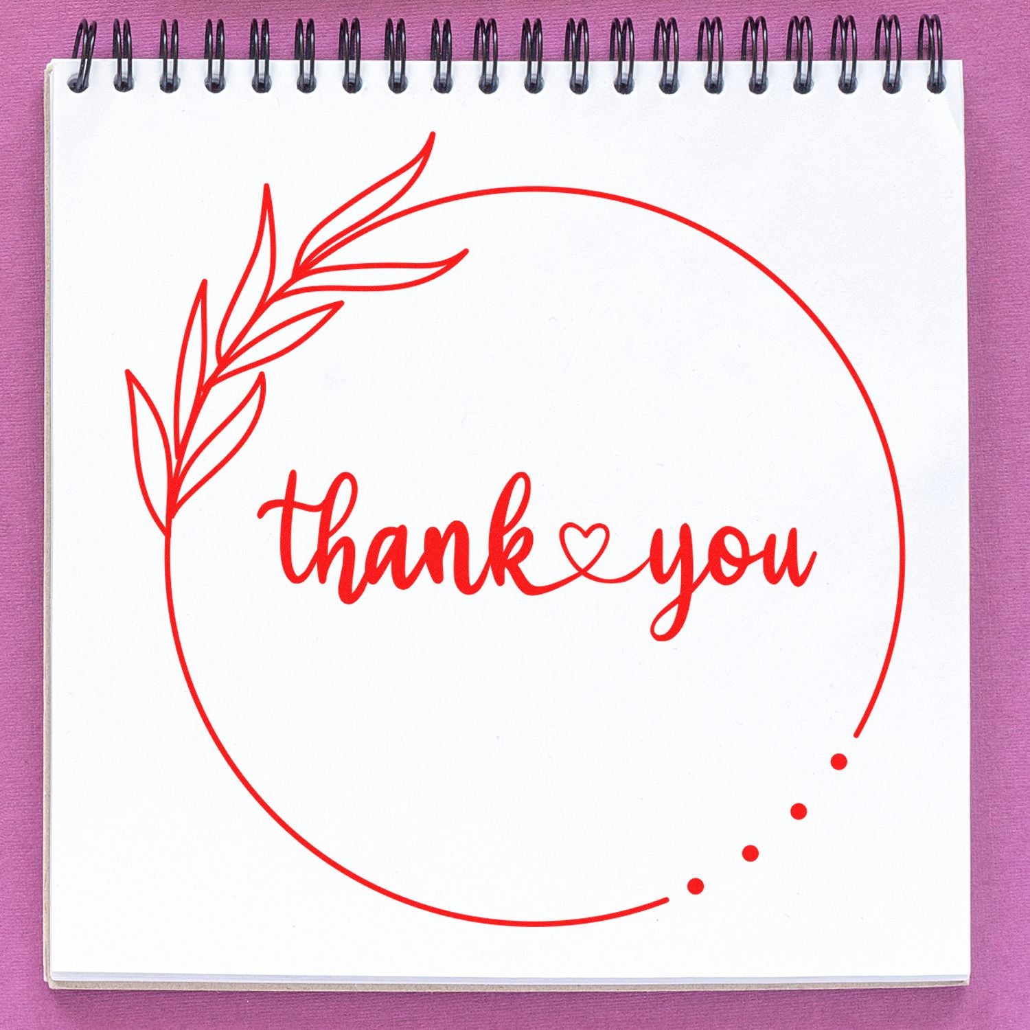 Red thank you stamp with leaf wreath and heart, forming a circular seal on a white spiral notebook over a purple background.