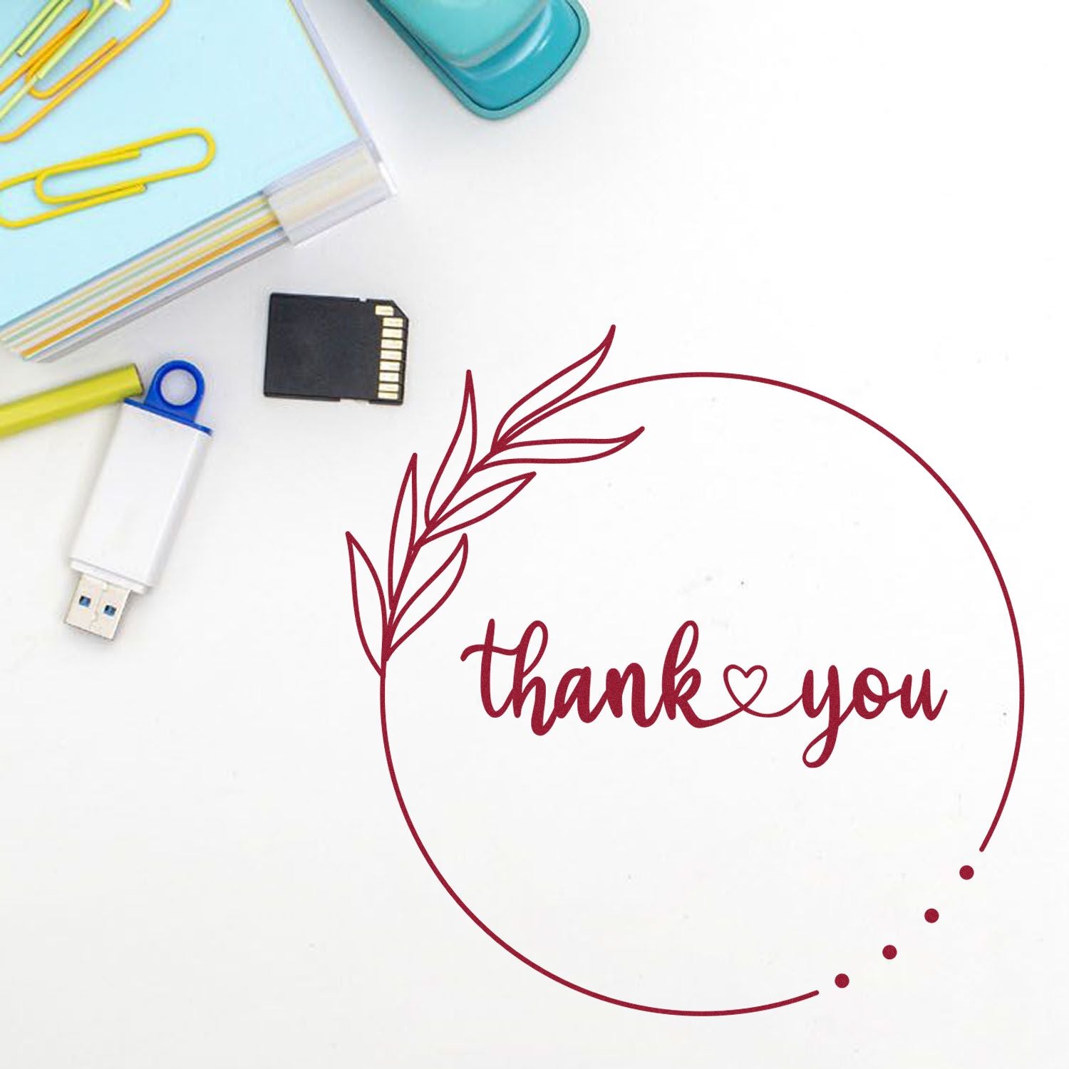 Burgundy thank you stamp seal with leafy wreath on white background, beside pastel office supplies and a memory card.