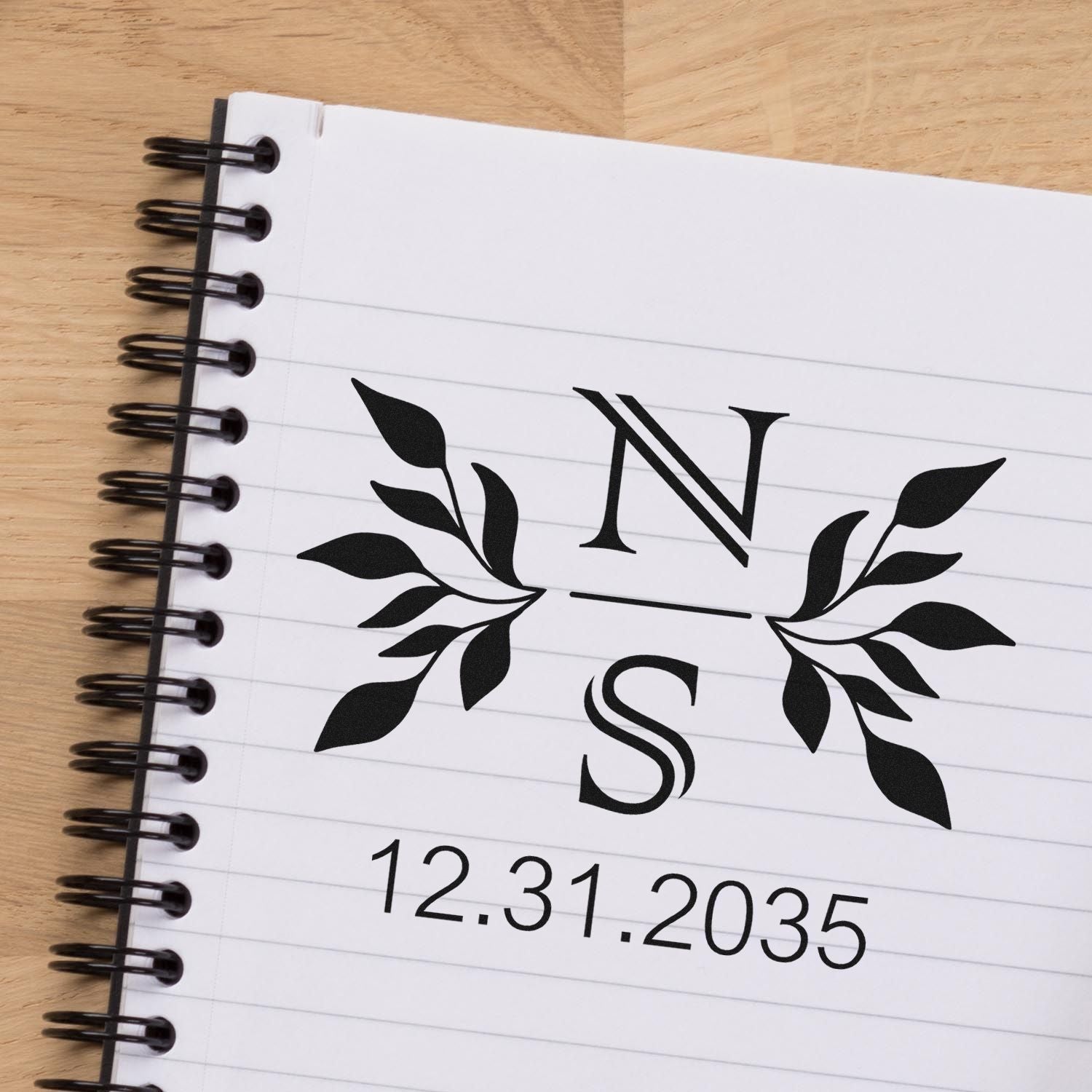 Black monogram stamp seal with leafy branches and date 12.31.2035 on a white lined spiral notebook.