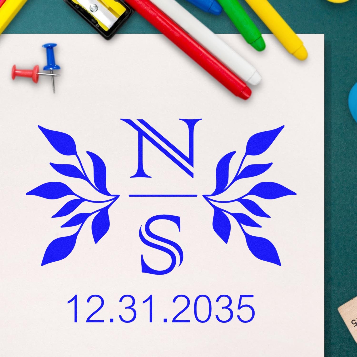Blue custom seal stamp with NS monogram, leafy wreath, and 12.31.2035 date on white paper, with colorful markers and push pins.