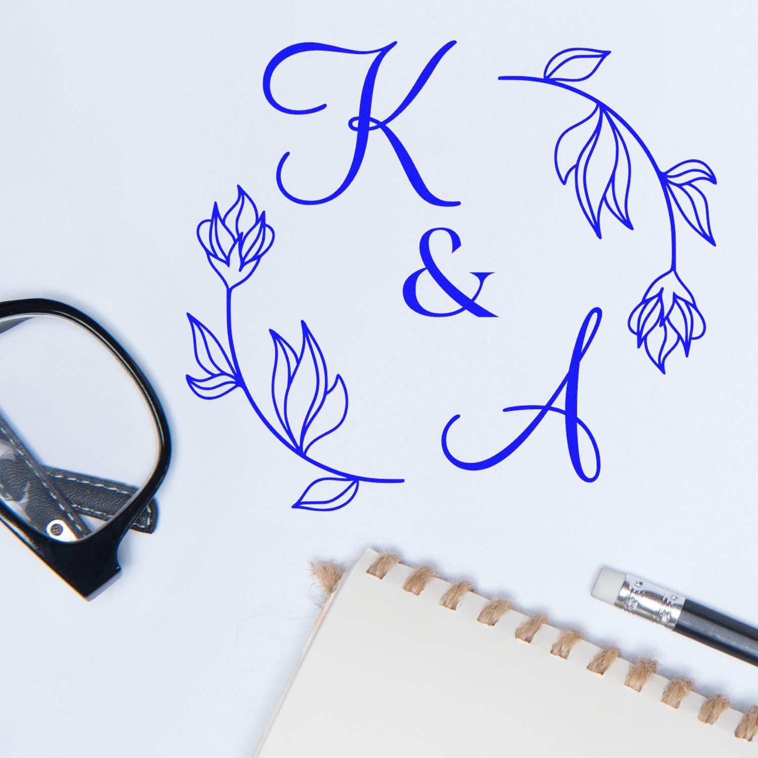 Blue K&A monogram stamp seal with floral wreath, shown with stamp tools and a notebook on white.