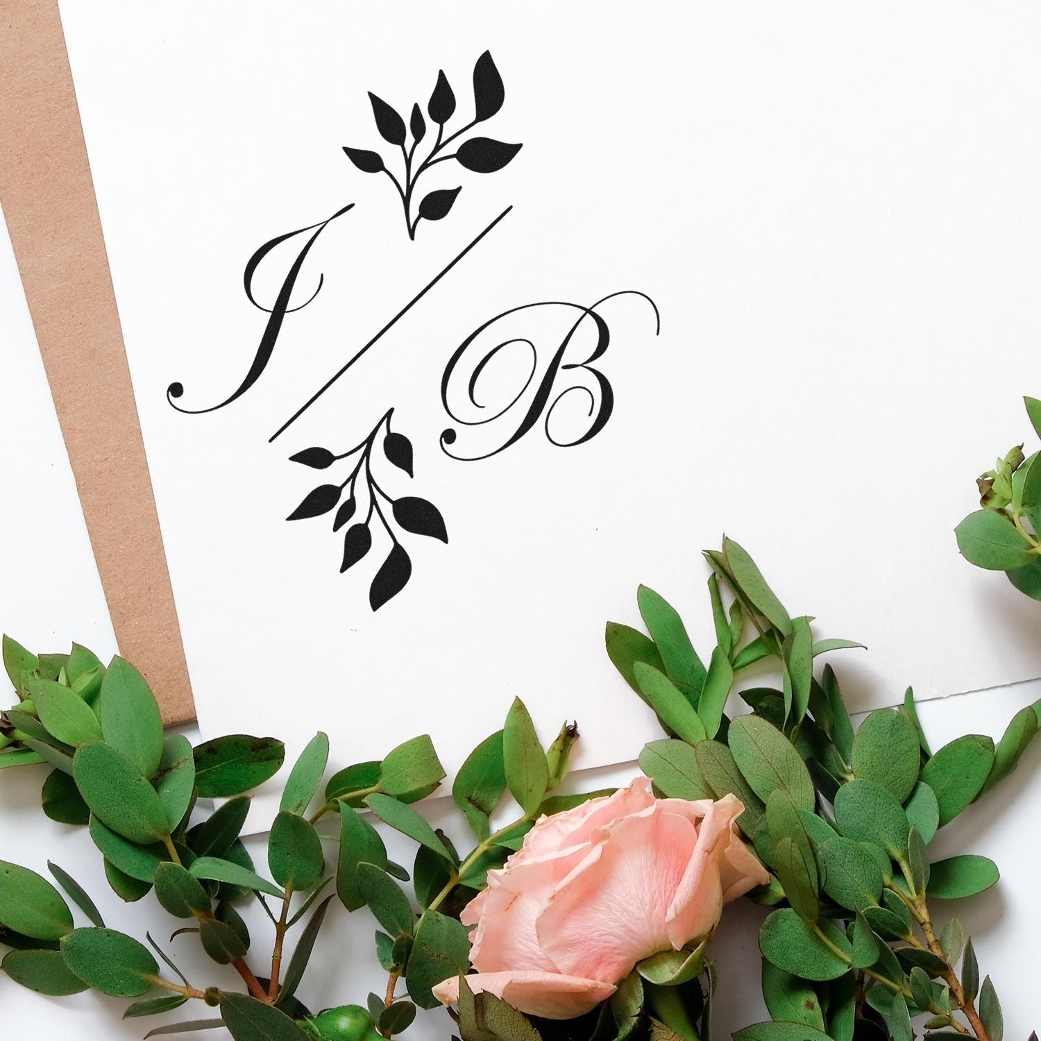 Black monogram stamp and seal design with leafy accents on white paper, surrounded by green foliage and a blush pink rose.