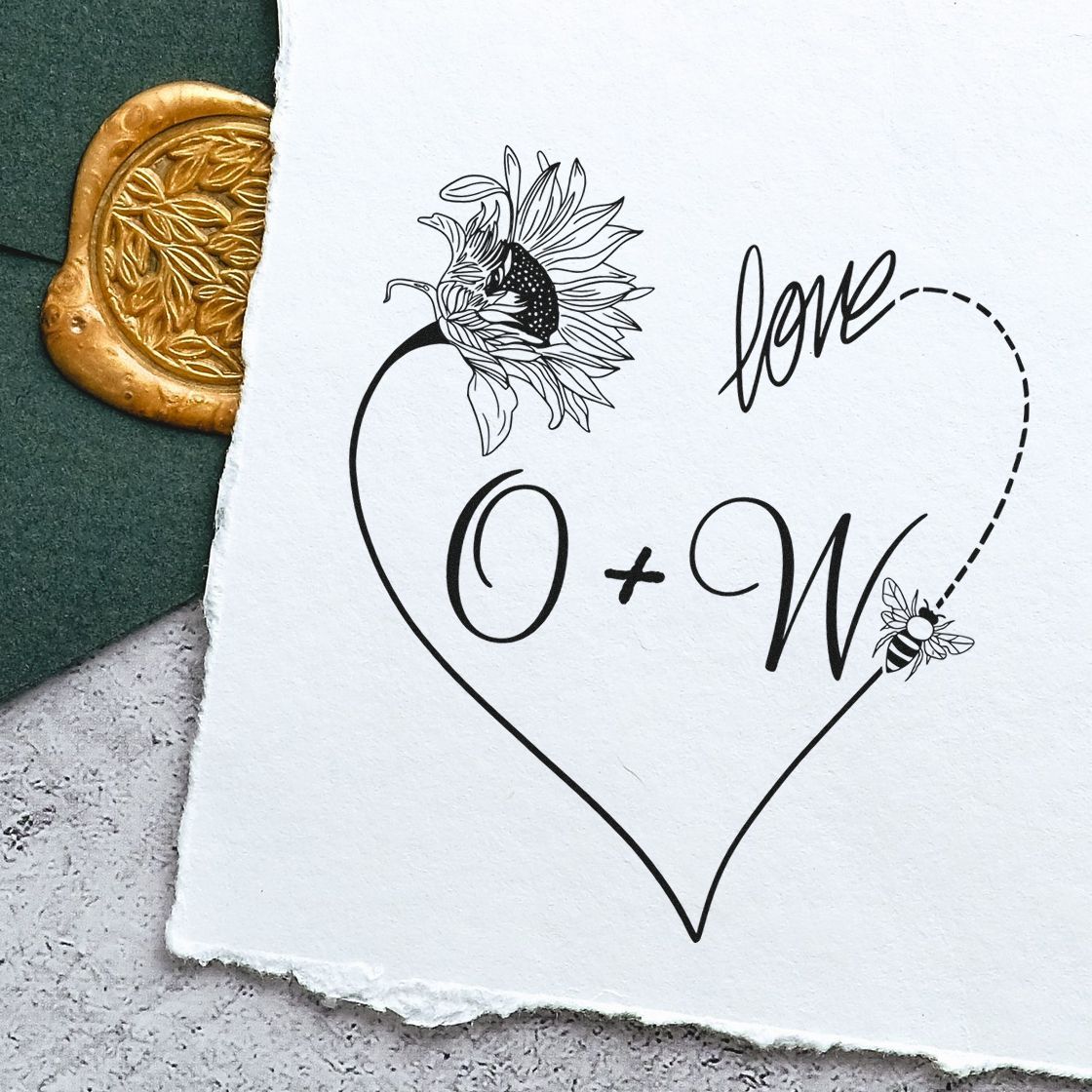Black heart-shaped stamp design with sunflower, bee, and love O+W on white paper beside a gold wax seal.