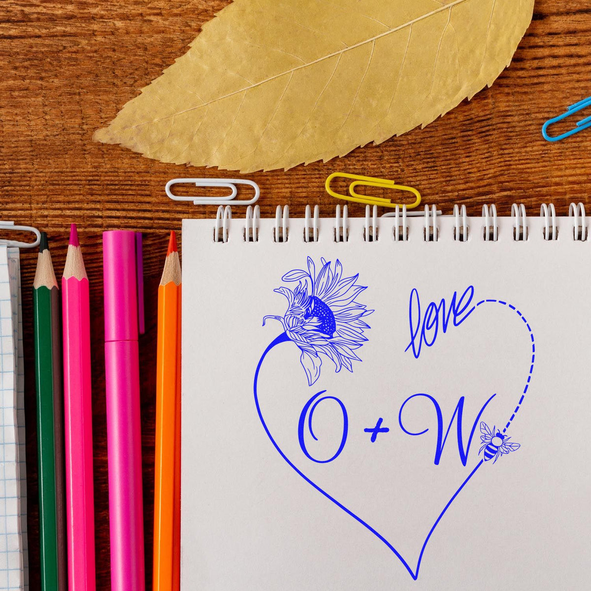 Blue heart-shaped love stamp with sunflower and bee seal on a white notebook, beside colorful pencils on a wood table.