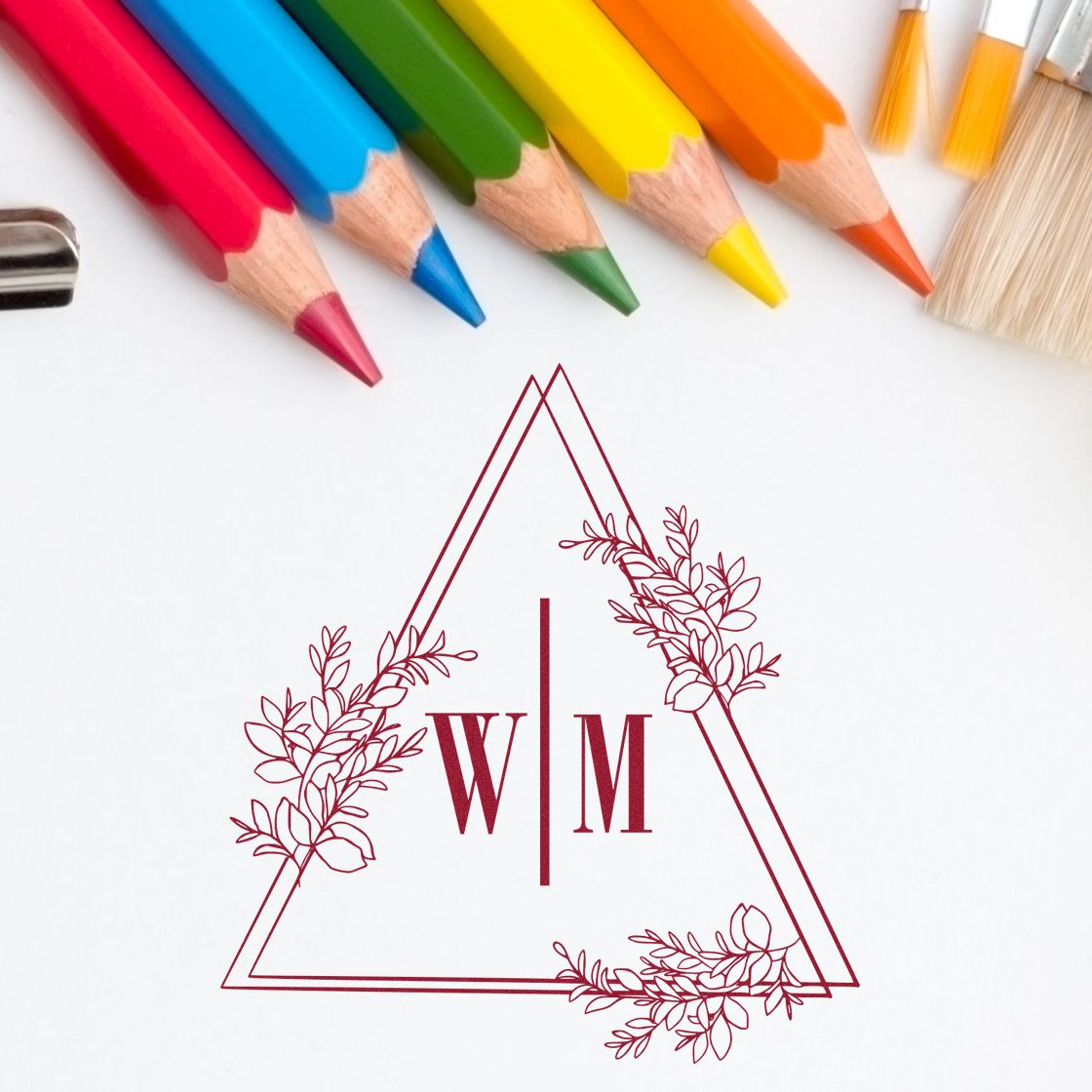 Custom burgundy floral monogram stamp seal with triangle outline and W|M initials beneath colorful pencils.
