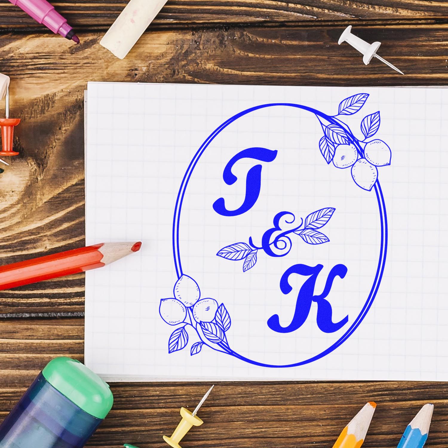 Blue monogram stamp seal with floral lemon accents on white paper, surrounded by colored pencils and push pins on a rustic wood desk.