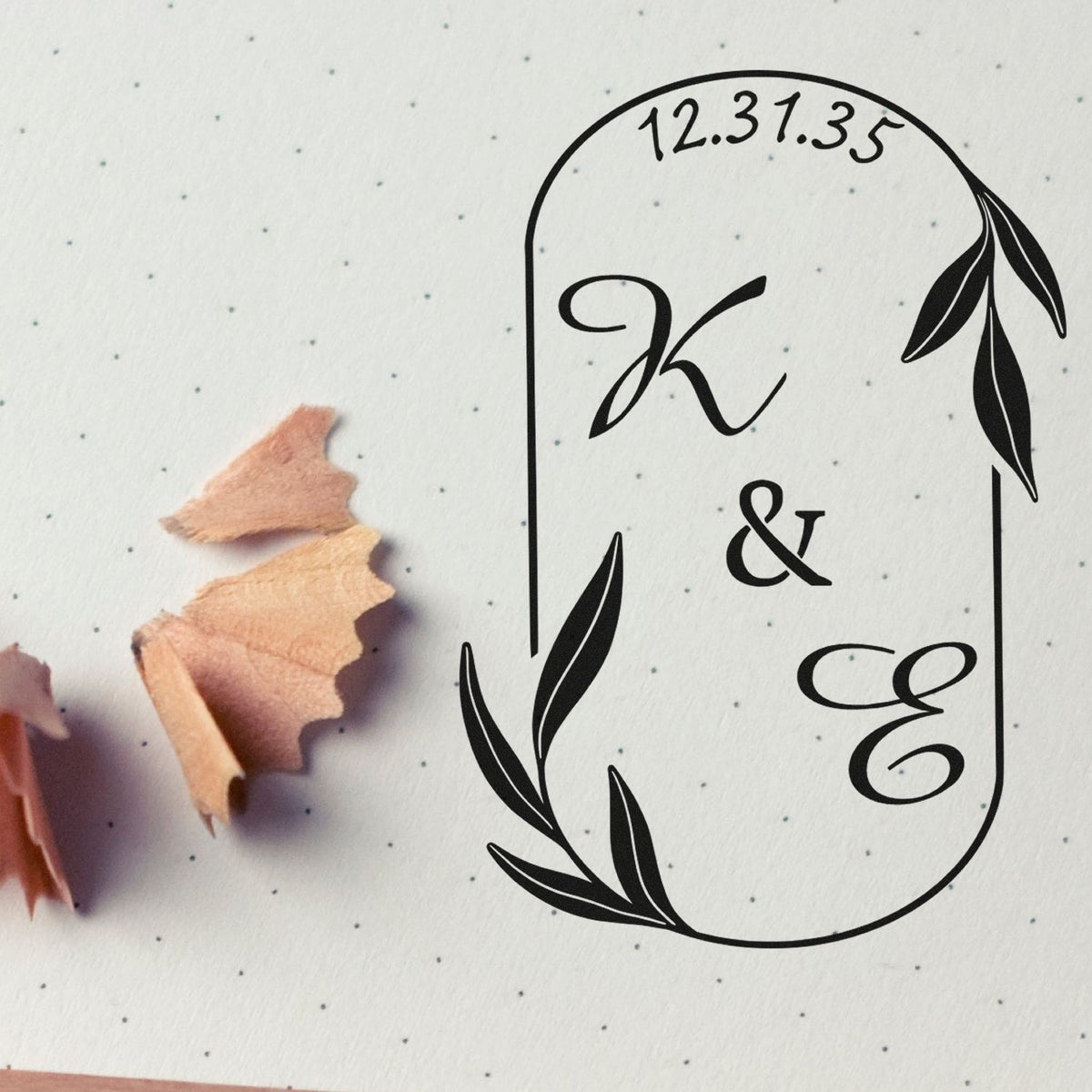 Black monogram stamp and seal design with leafy border on white dotted paper, beside pink pencil shavings.