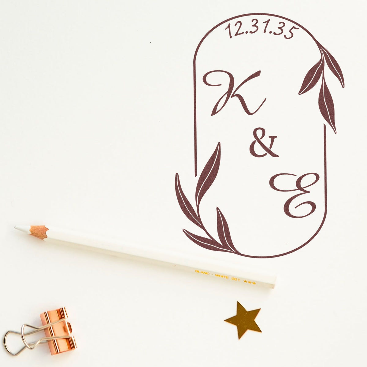Burgundy custom monogram stamp and seal with leafy arch, date 12.31.35, on white paper with pencil, binder clip, and gold star.