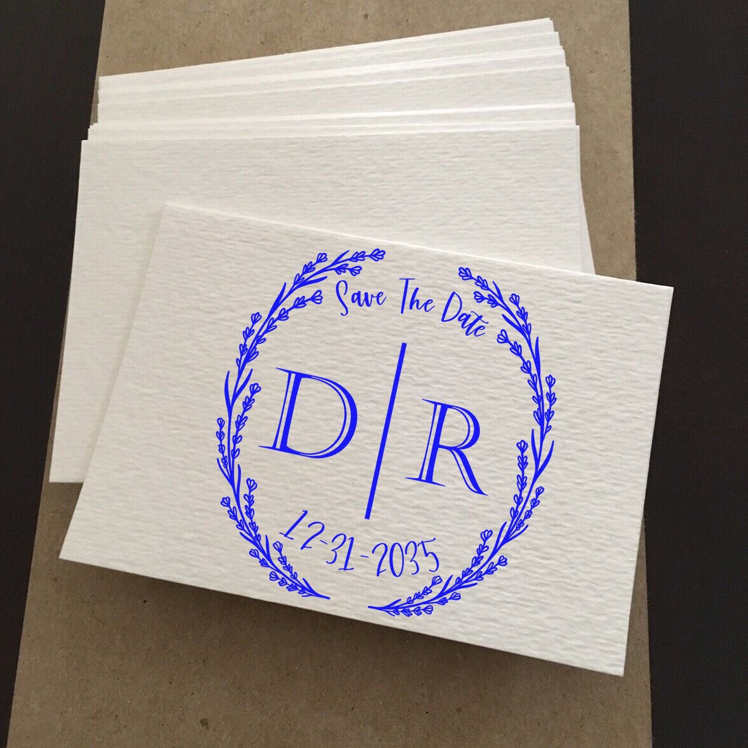 Blue Save The Date stamp and seal design with floral wreath, D/R monogram, and 12-31-2035 on white textured envelopes.