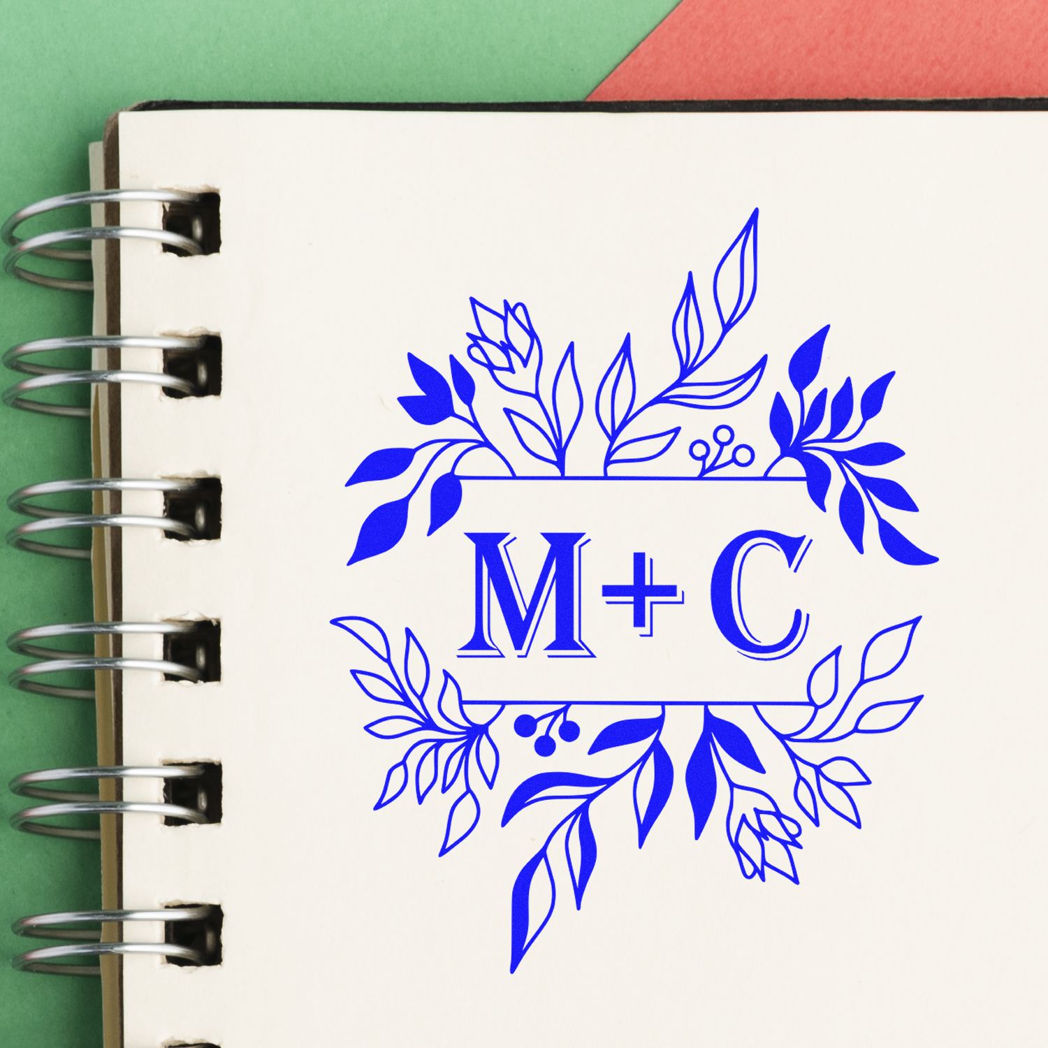 Custom blue floral stamp seal with M+C on a cream spiral notebook, on a green and red background.