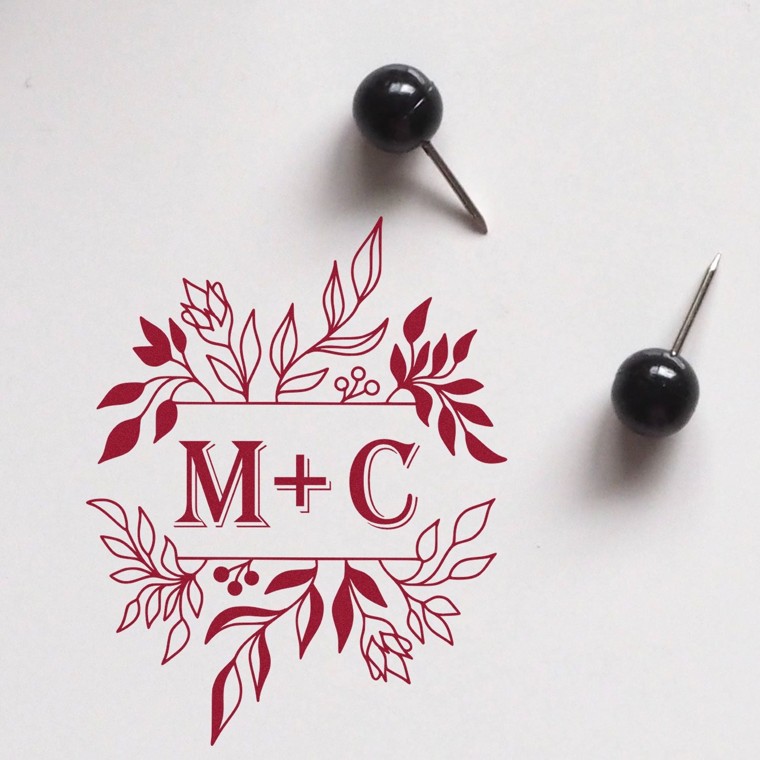 Red floral M+C stamp and seal design with two black pushpin tools on a white background.