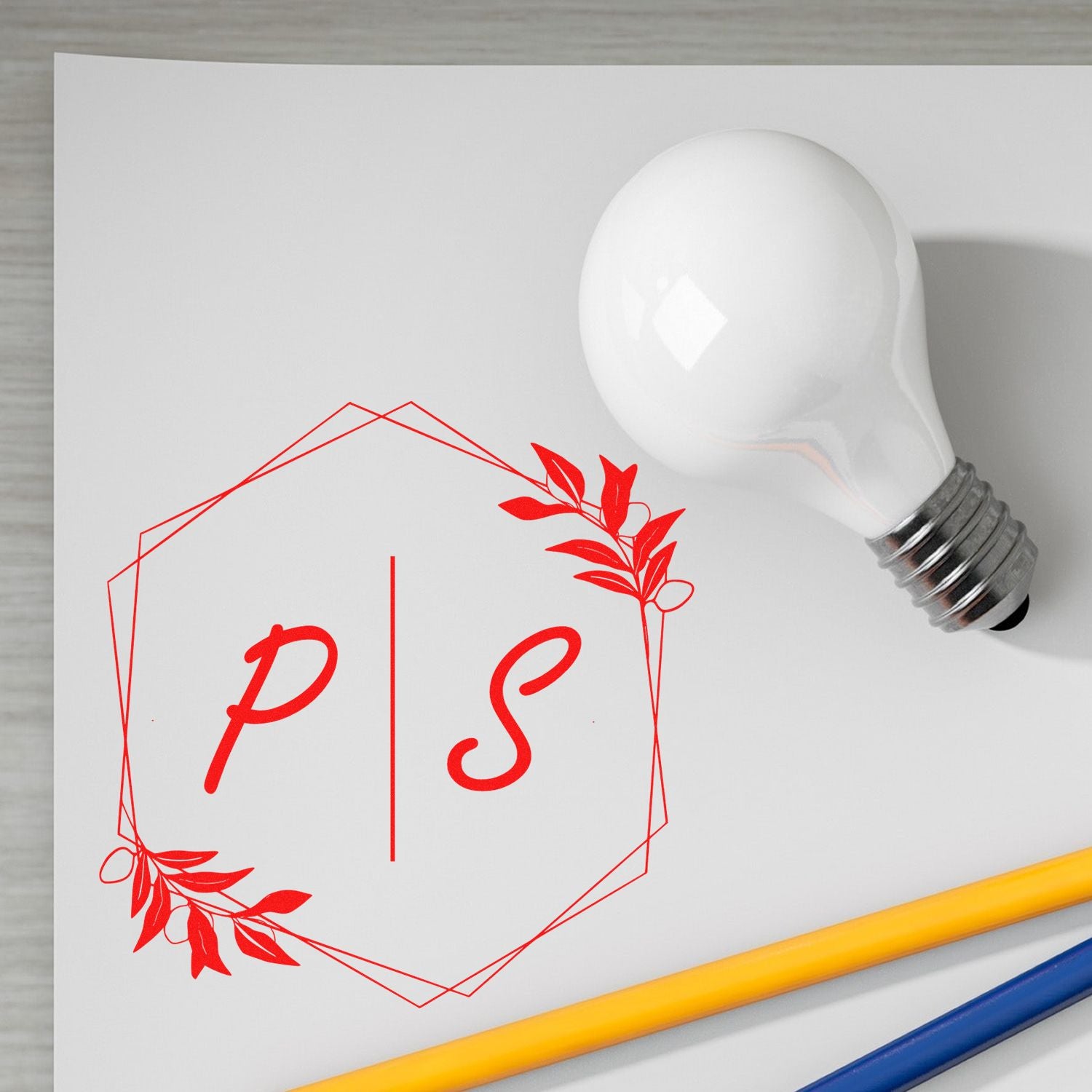 Red PS stamp and seal logo in a floral geometric frame on white paper, with a white bulb and yellow-blue pencils.
