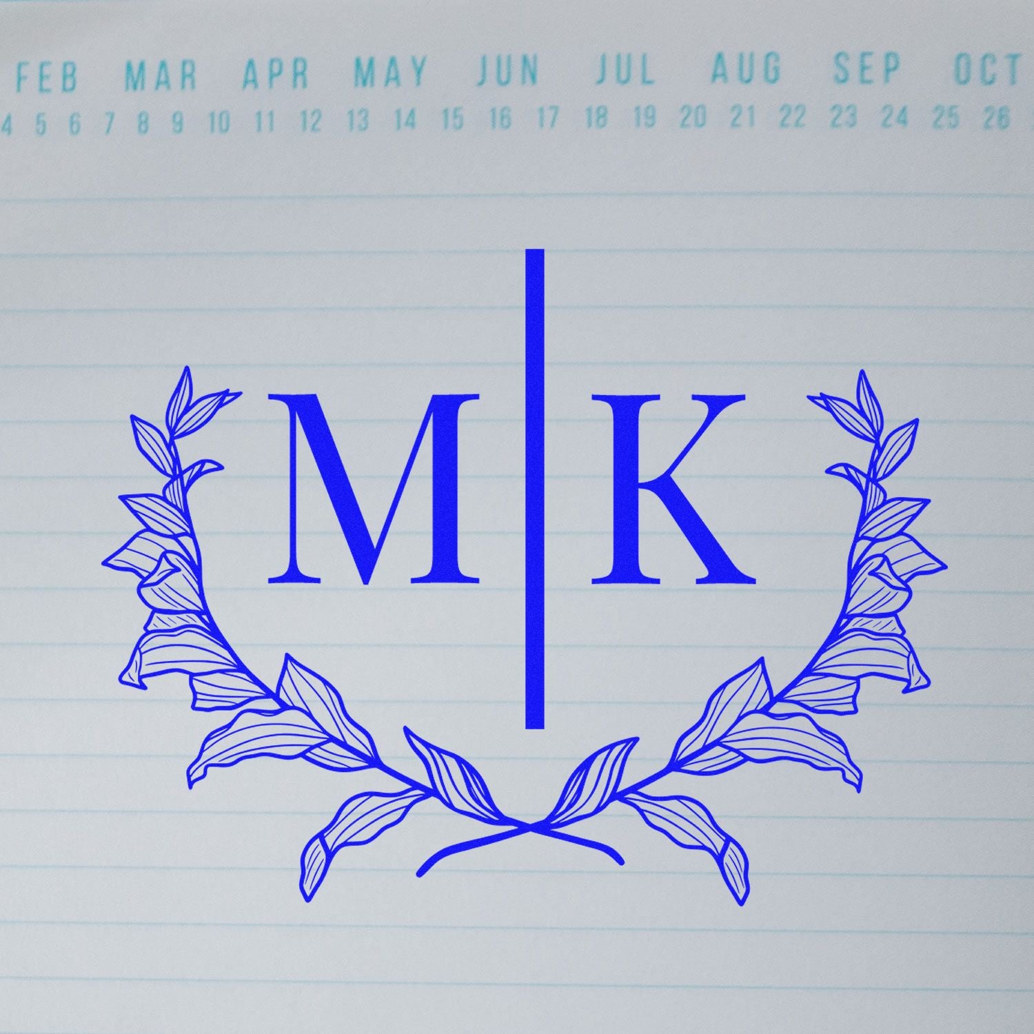 Blue custom monogram stamp seal with M|K in serif lettering, framed by a leafy wreath on lined paper.