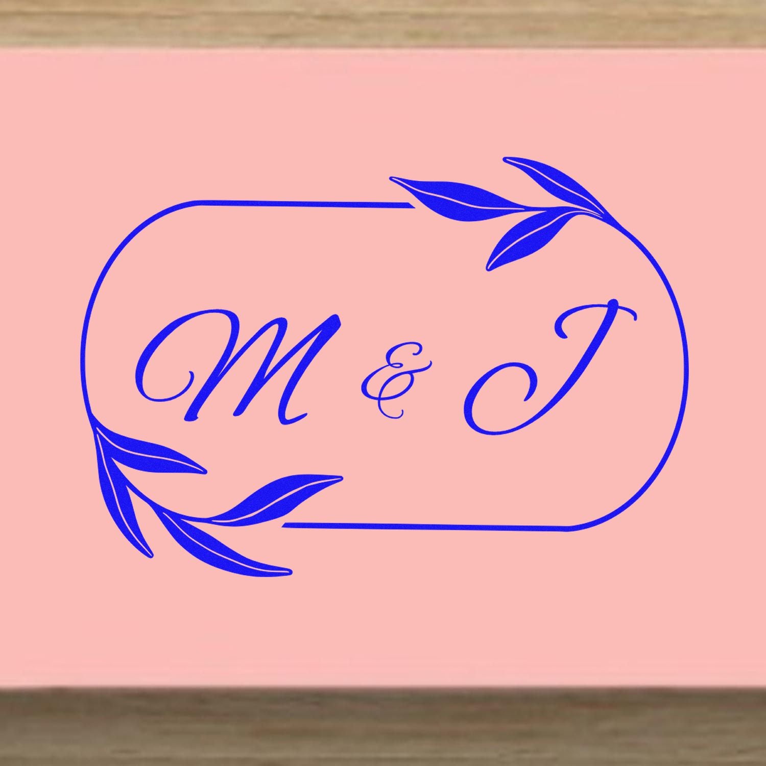 Pink custom stamp seal with royal blue M & J monogram and leafy border accents.