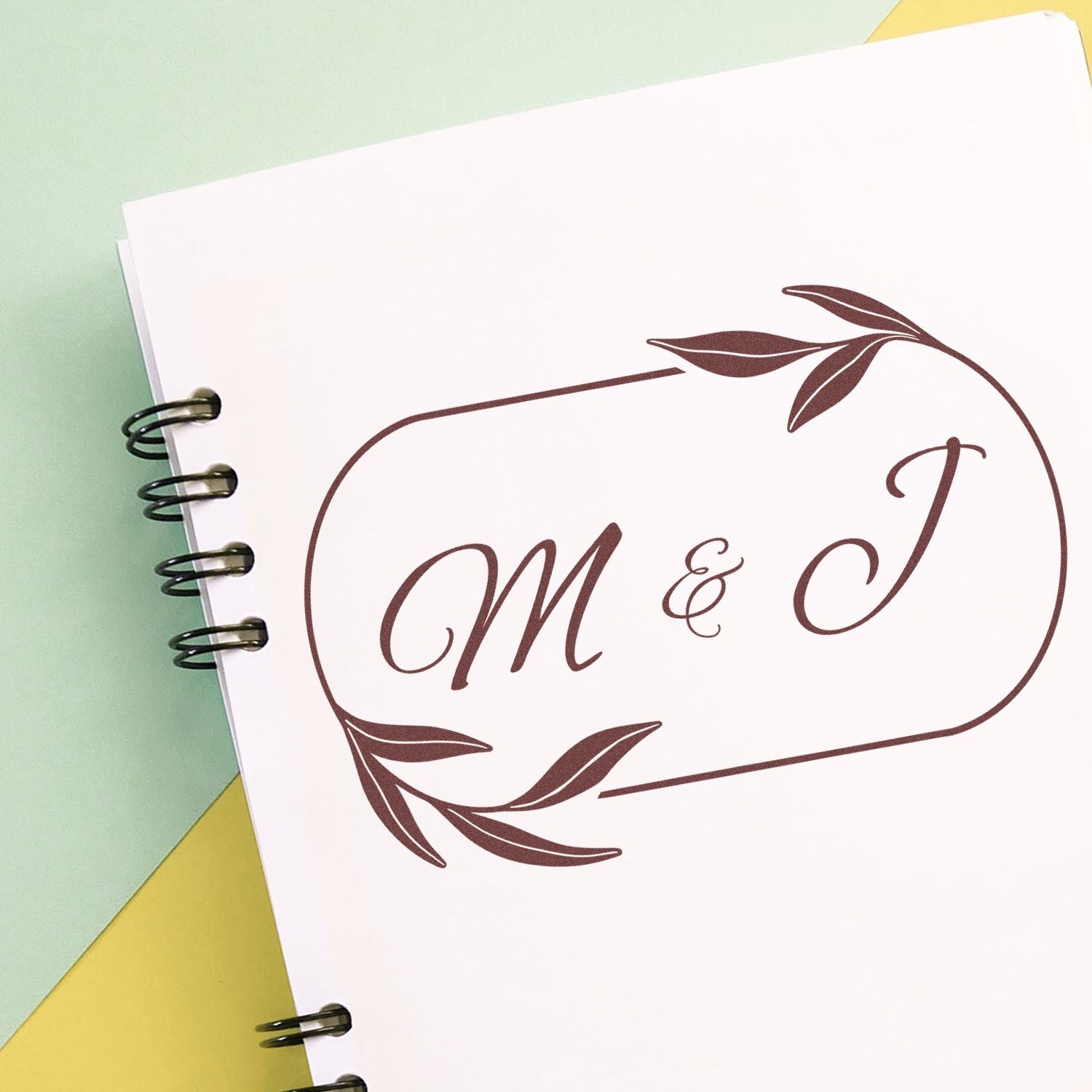 Brown monogram stamp seal with M & J and leafy wreath on white spiral-bound paper against pastel green and yellow background.