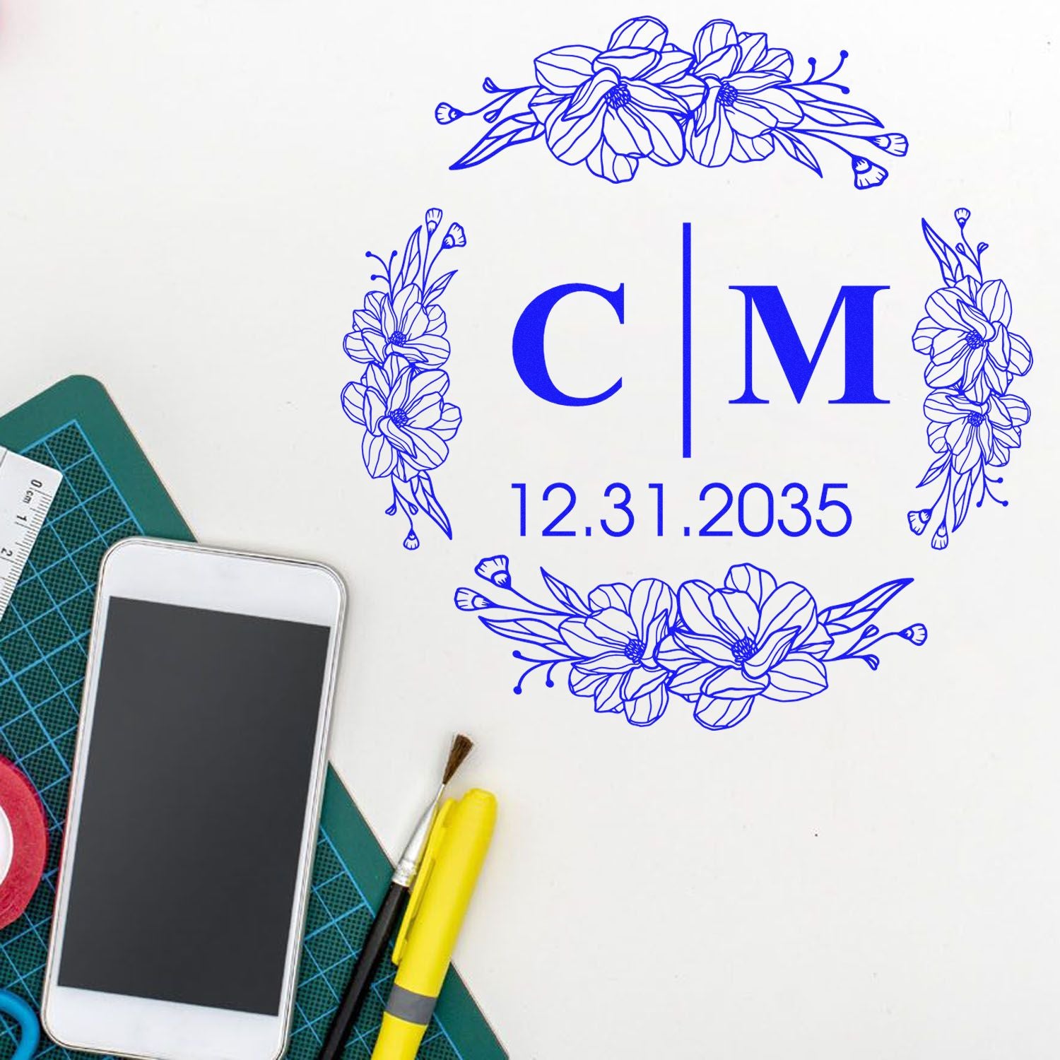 Blue floral stamp and seal design with C|M monogram and 12.31.2035 date, shown on a craft desk with tools.