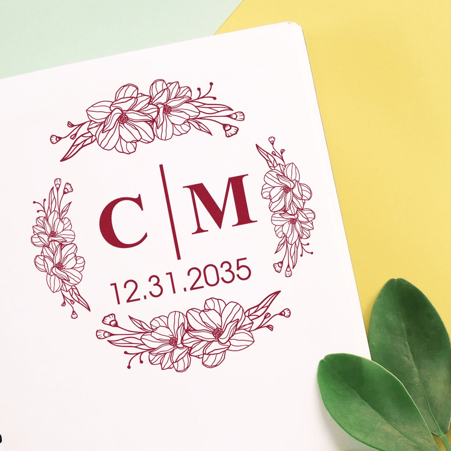 Maroon floral monogram stamp seal with C|M and 12.31.2035 on a white card, against yellow and green background.