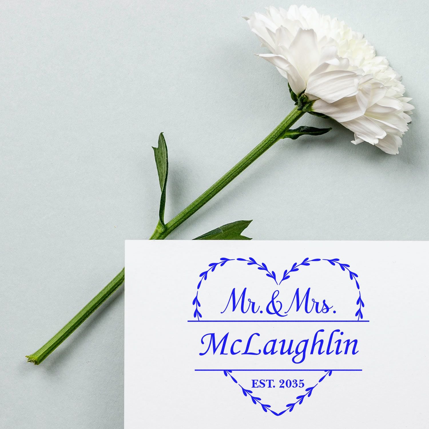 White flower beside blue custom wedding stamp seal reading Mr. & Mrs. McLaughlin, EST. 2035, in heart wreath design.