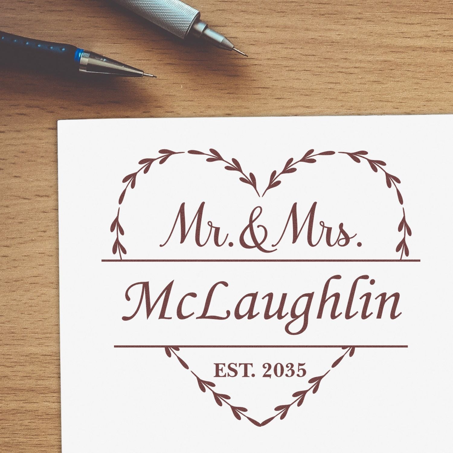 Burgundy custom wedding stamp and seal with heart wreath, Mr. & Mrs. McLaughlin, EST. 2035, on white paper beside pens.