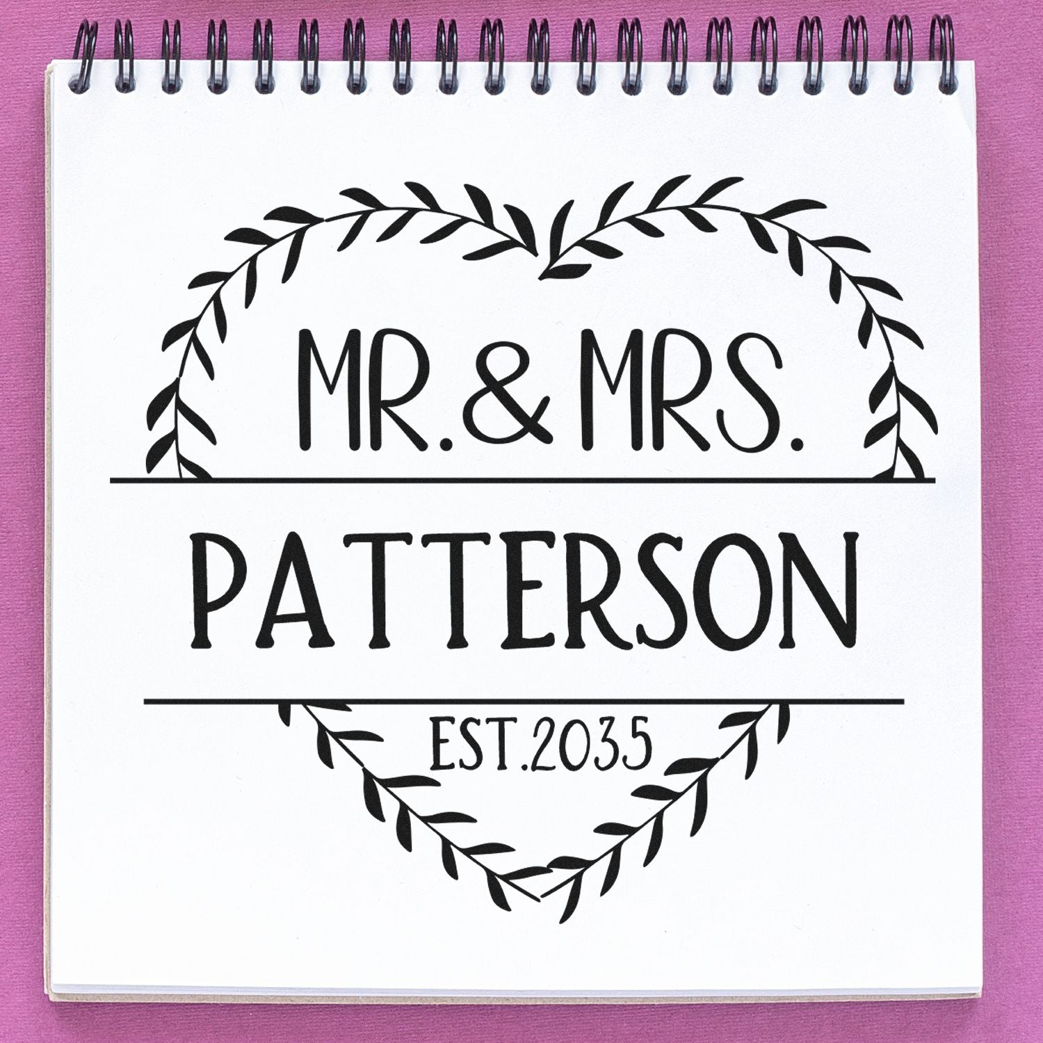 Custom wedding stamp and seal with leafy heart frame, black MR. & MRS. PATTERSON, EST. 2035 on white notebook, pink background.