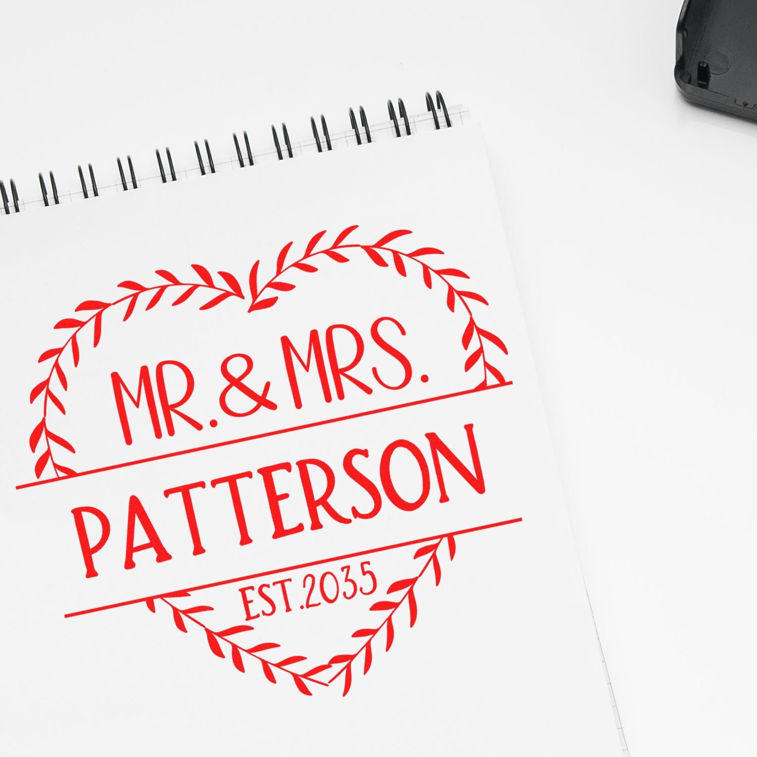 Red custom stamp seal on white spiral notepad, reading Mr. & Mrs. Patterson with leafy wreath and Est. 2035.