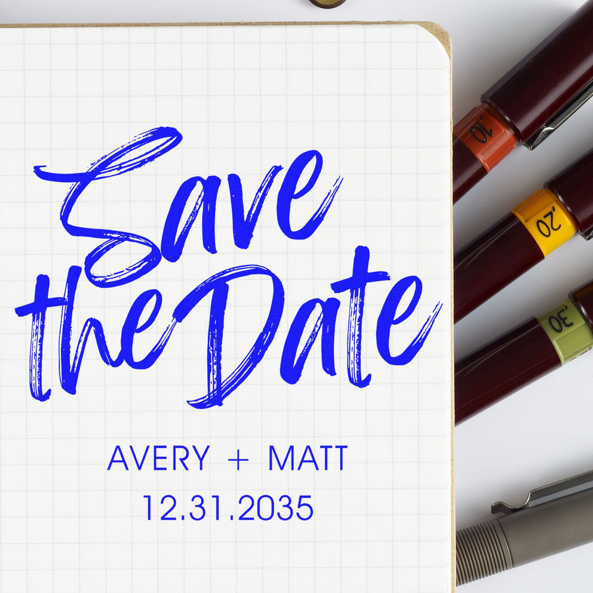 Blue Save the Date wedding stamp on grid paper with Avery + Matt 12.31.2035 and colored stamp and seal pens.