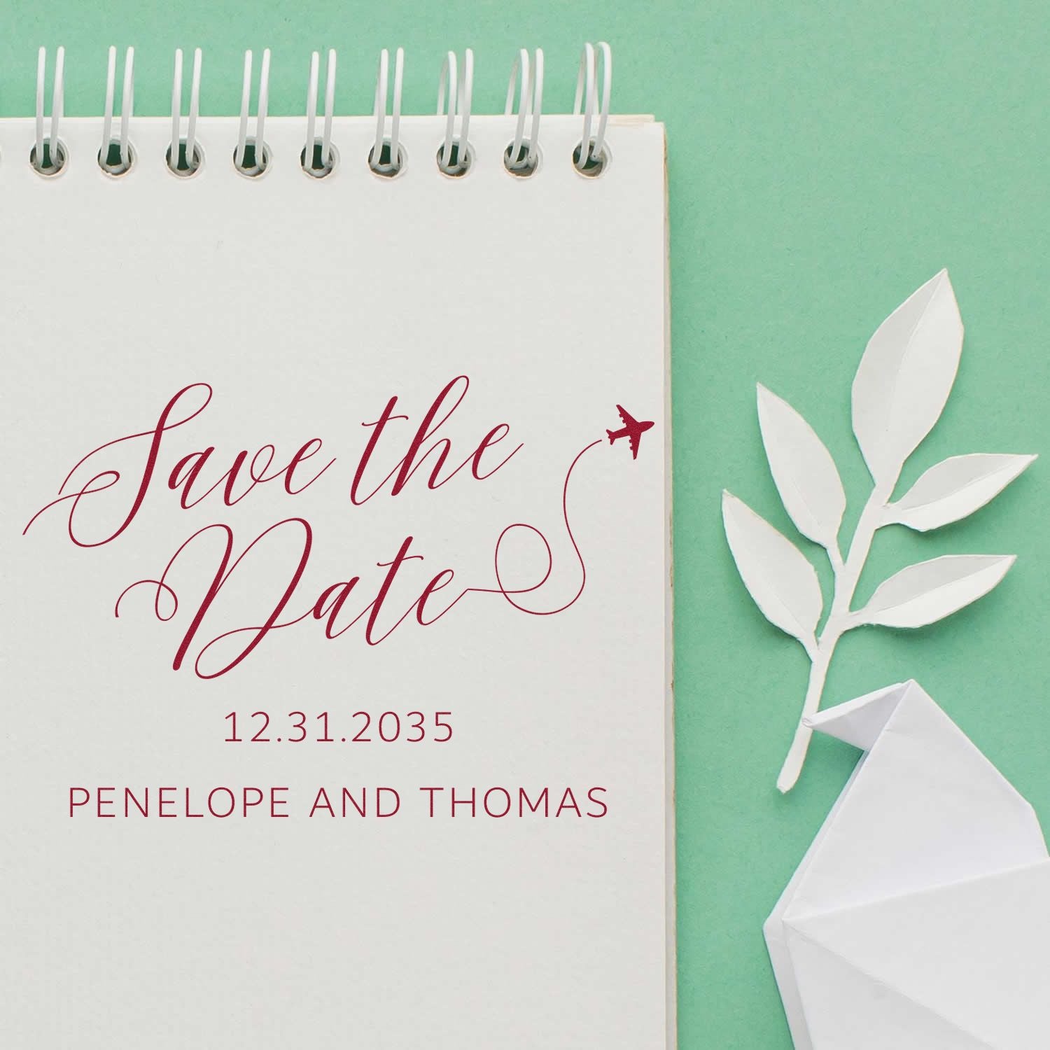 Burgundy Save the Date stamp on a white spiral notebook, with date and names, beside a white paper leaf and envelope seal on mint green.