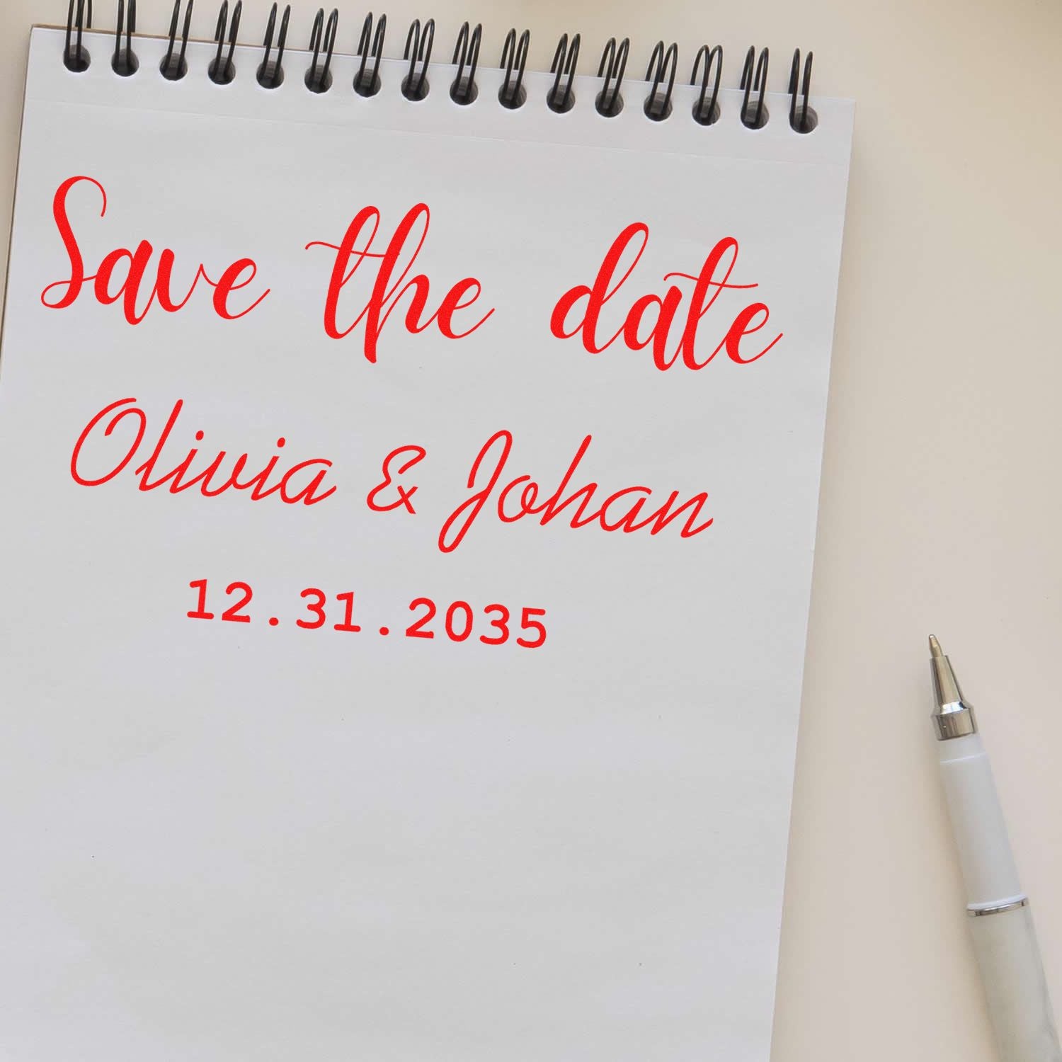 Red calligraphy save-the-date stamp seal on white spiral notepad, reading Olivia & Johan 12.31.2035, with pen.