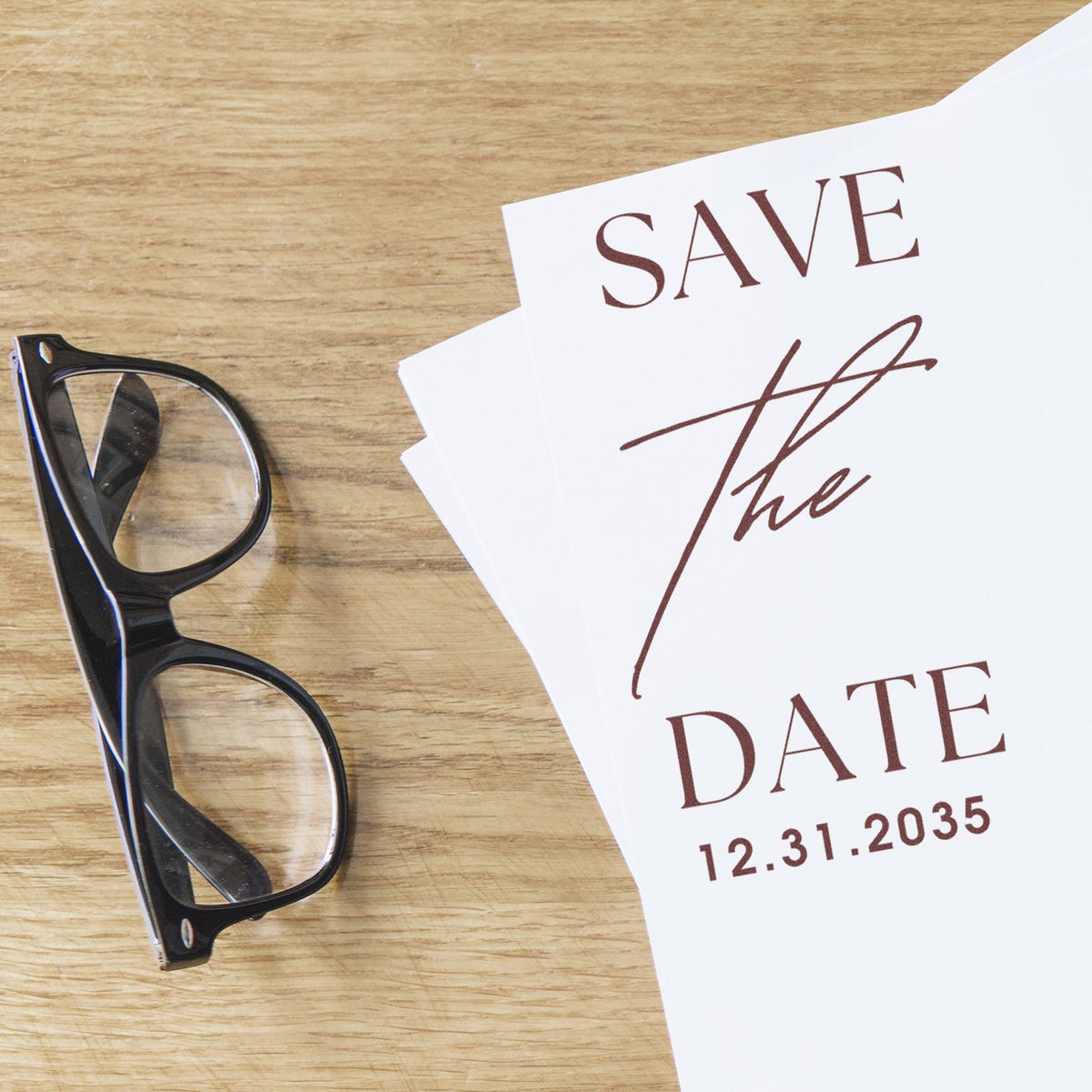Black glasses beside white save-the-date stamp and seal cards with brown text on wood tabletop.