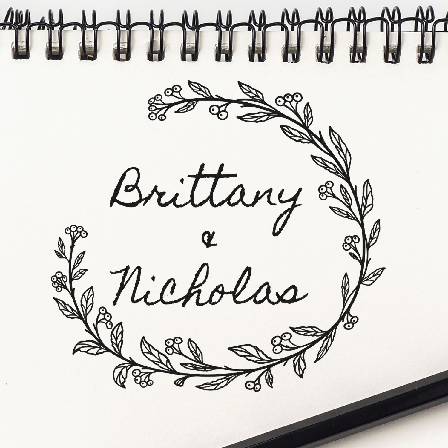 Black floral wreath stamp and seal design with Brittany & Nicholas on a white spiral-bound page.