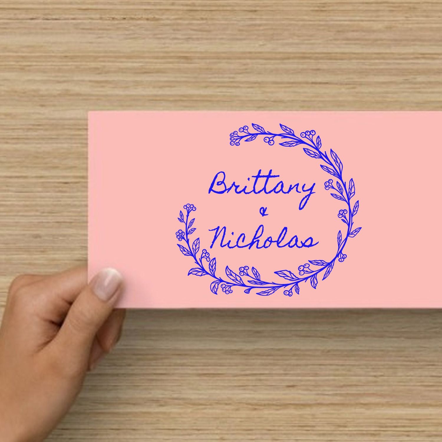 Pink custom stamp seal with blue floral wreath and Brittany & Nicholas text, held over a wood table.