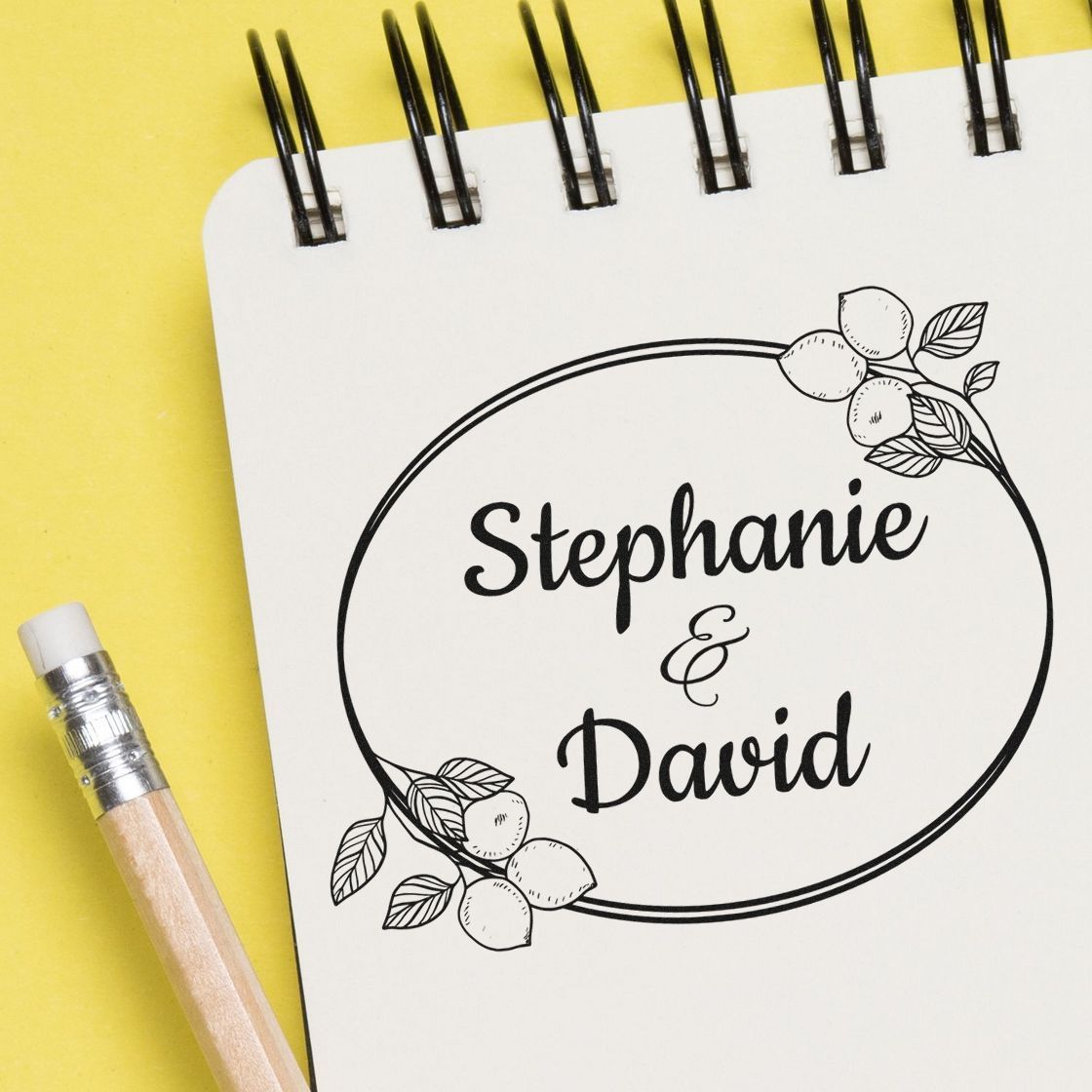 Custom wedding stamp and seal design with black Stephanie & David lettering, floral accents, on a white notebook over yellow background.