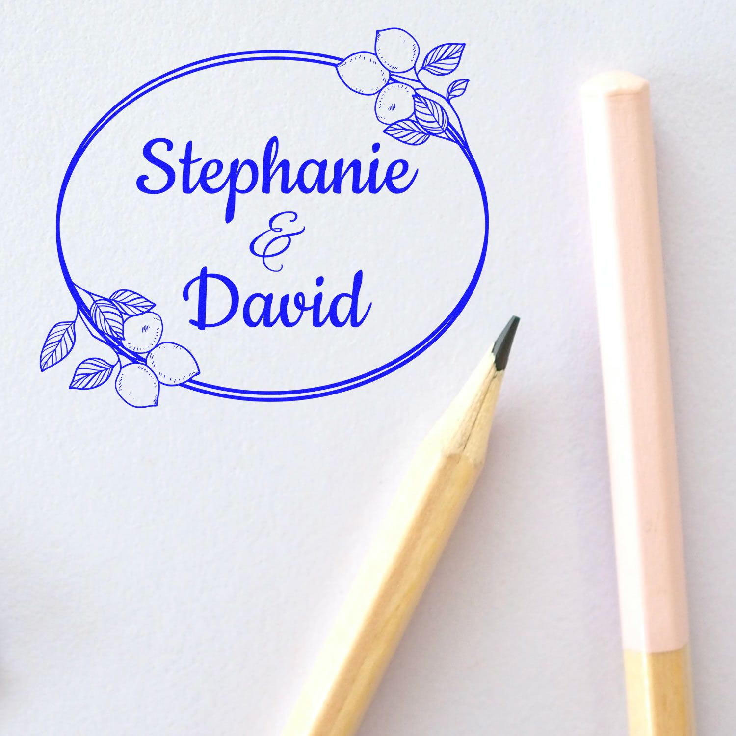 Blue oval custom stamp and seal for Stephanie & David, with lemon floral accents on white paper beside pastel pencils.