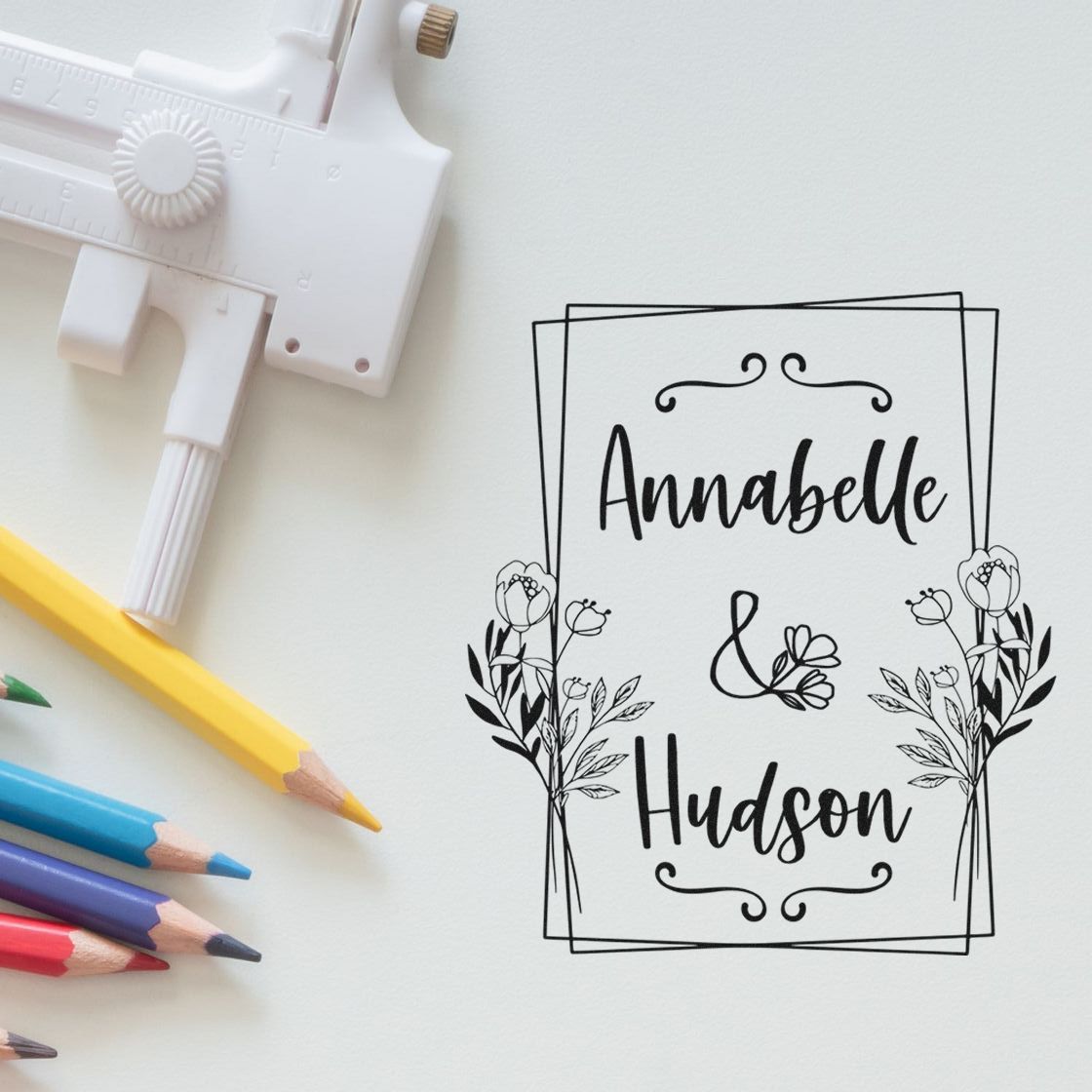 White custom stamp tool with colorful pencils beside black floral seal for Annabelle & Hudson on white paper.