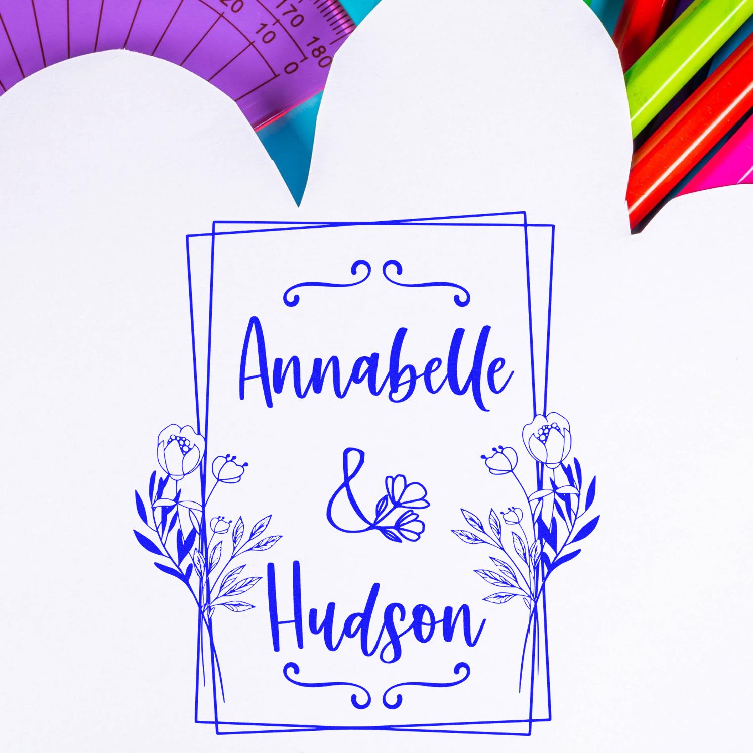 Blue custom wedding stamp seal with Annabelle & Hudson, floral border, on white paper with colorful rulers and pens.