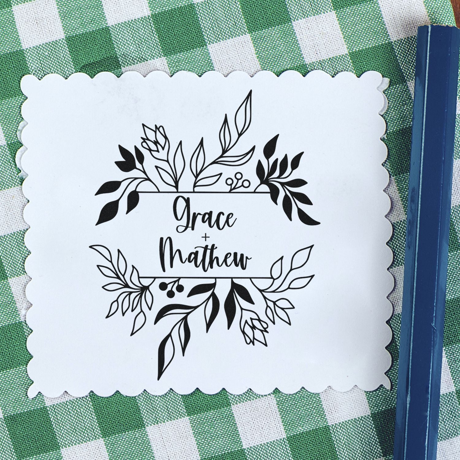 White scalloped stamp seal with black floral Grace + Matthew wedding design on green gingham, beside a blue stamping tool.