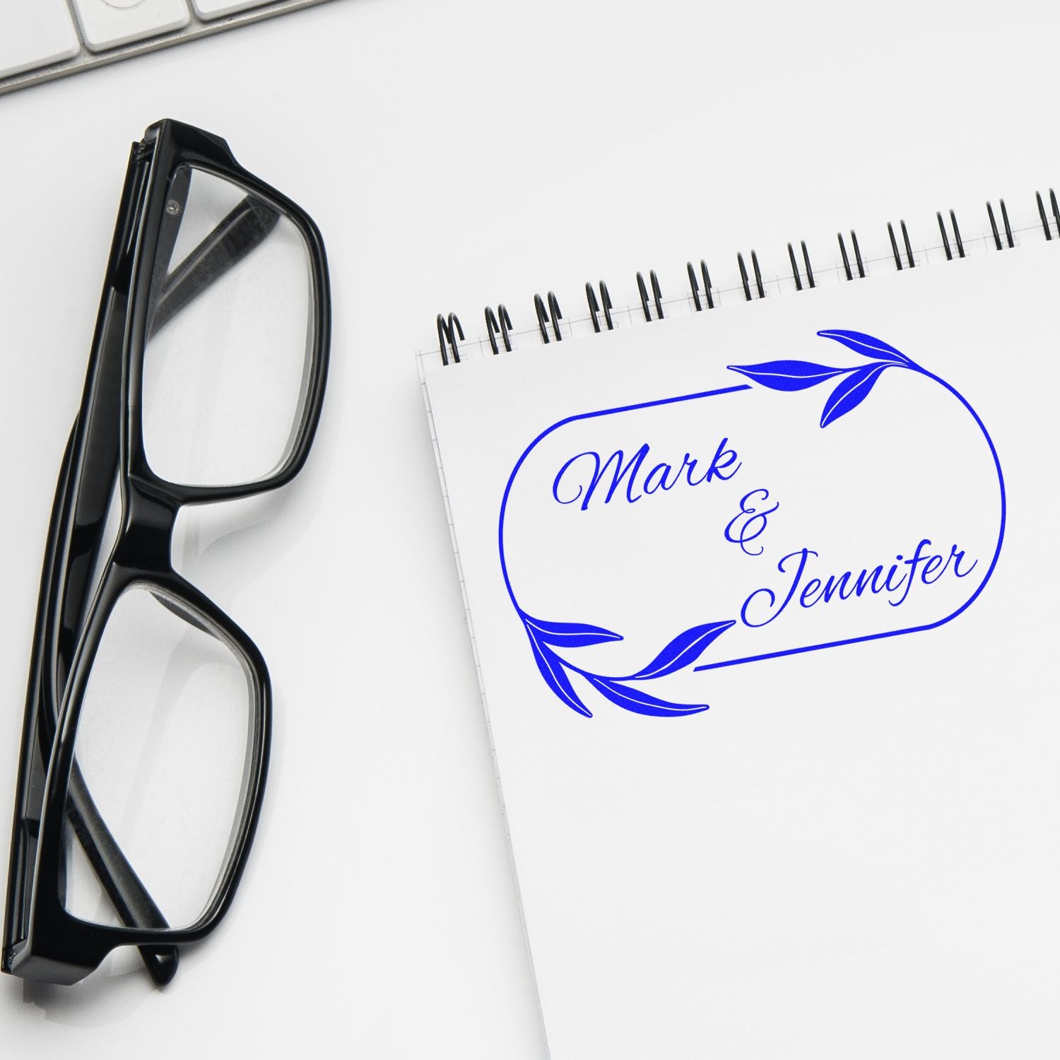 Black eyeglasses beside a white spiral notebook with a blue custom stamp and seal design reading Mark & Jennifer with leaf accents.