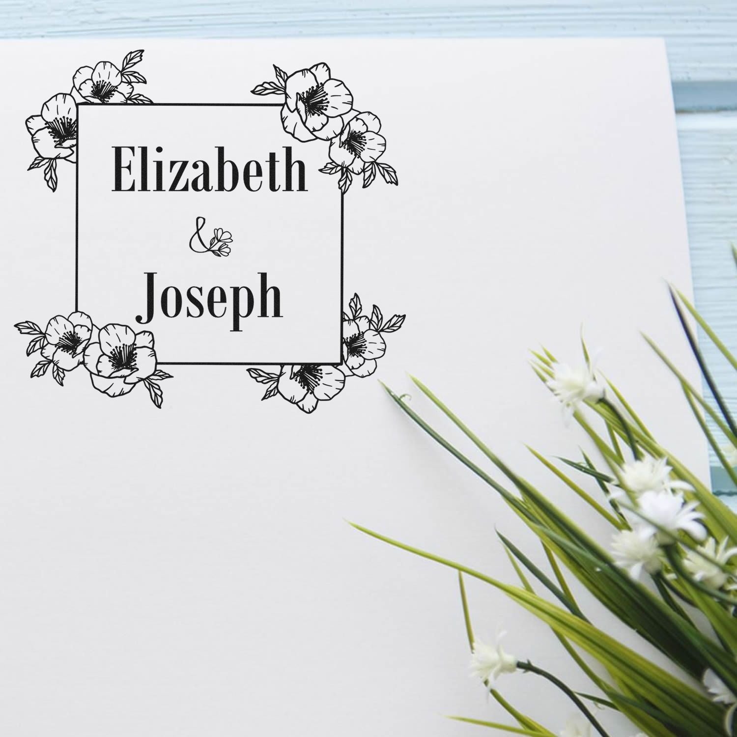 Black floral custom wedding stamp and seal with Elizabeth & Joseph on a white card, accented by green stems and white flowers.