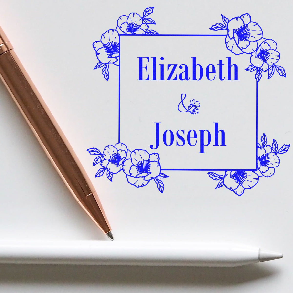 Blue floral custom stamp seal with Elizabeth & Joseph in a square frame, beside a rose-gold pen and white stylus.