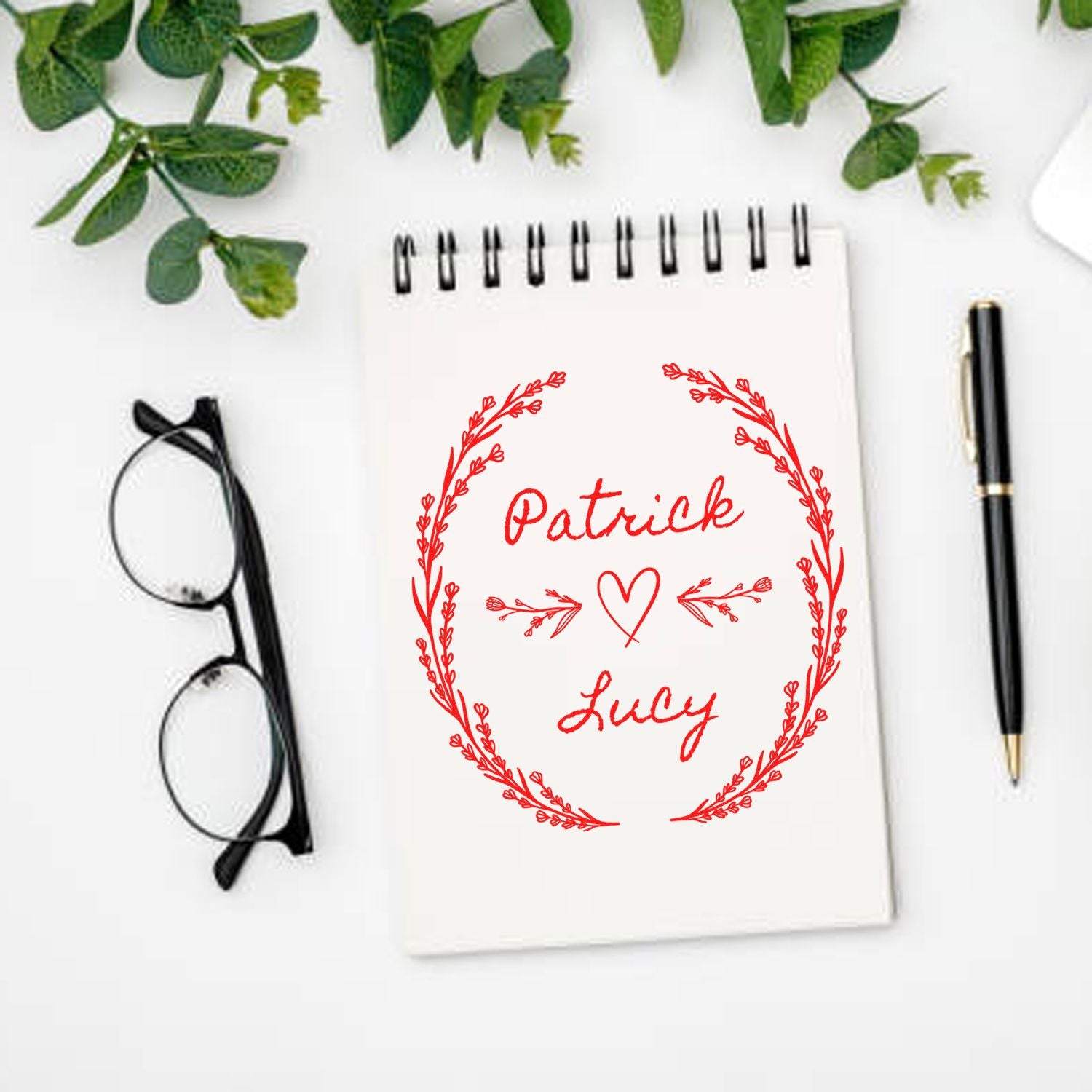 Red custom stamp seal with Patrick and Lucy, heart and laurel wreath on white notepad, beside glasses and a black pen.