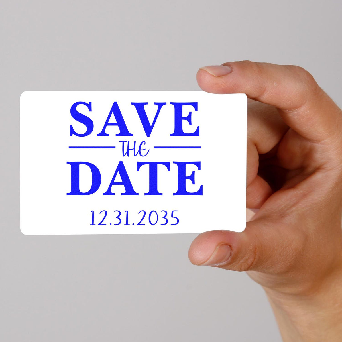 Hand holding a white save-the-date stamp and seal card with blue text and 12.31.2035 on a light gray background.