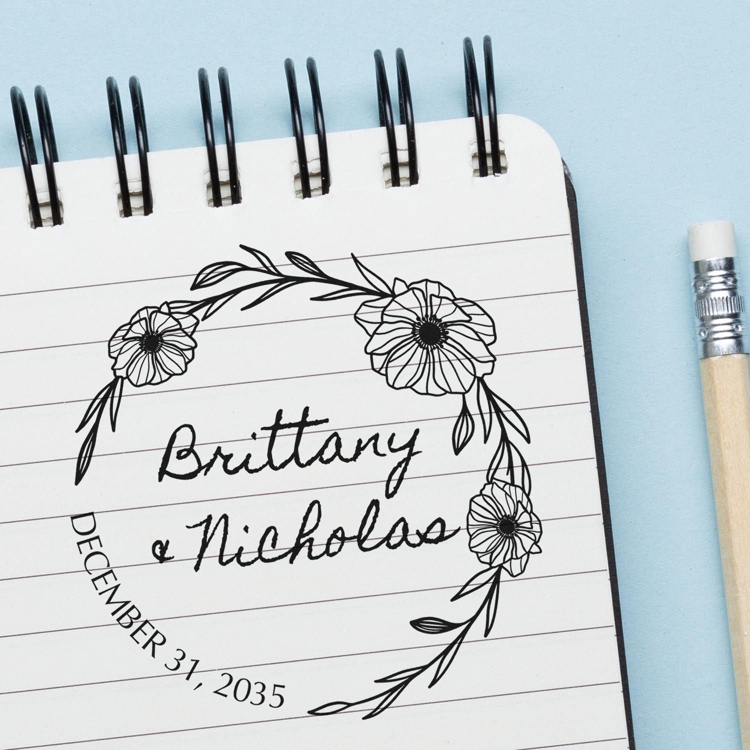 Custom floral stamp and seal on white spiral notebook, reading Brittany Nicholas and December 31, 2035, on blue background