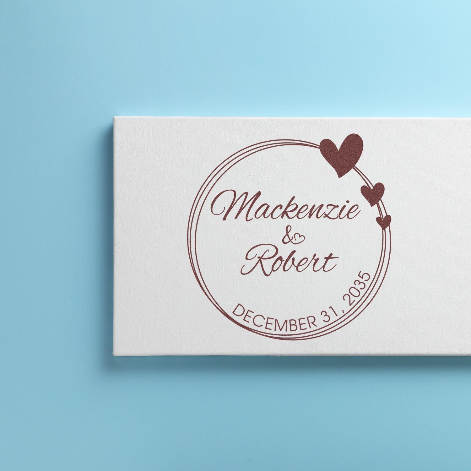 Custom burgundy wedding stamp and seal with hearts, Mackenzie & Robert, and December 31, 2035 on a white box over blue background.