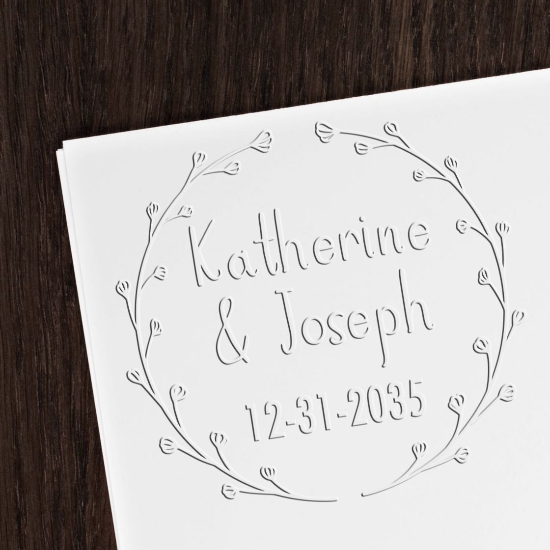 White embossed wedding stamp seal with floral wreath, Katherine & Joseph, and 12-31-2035 on dark wood.