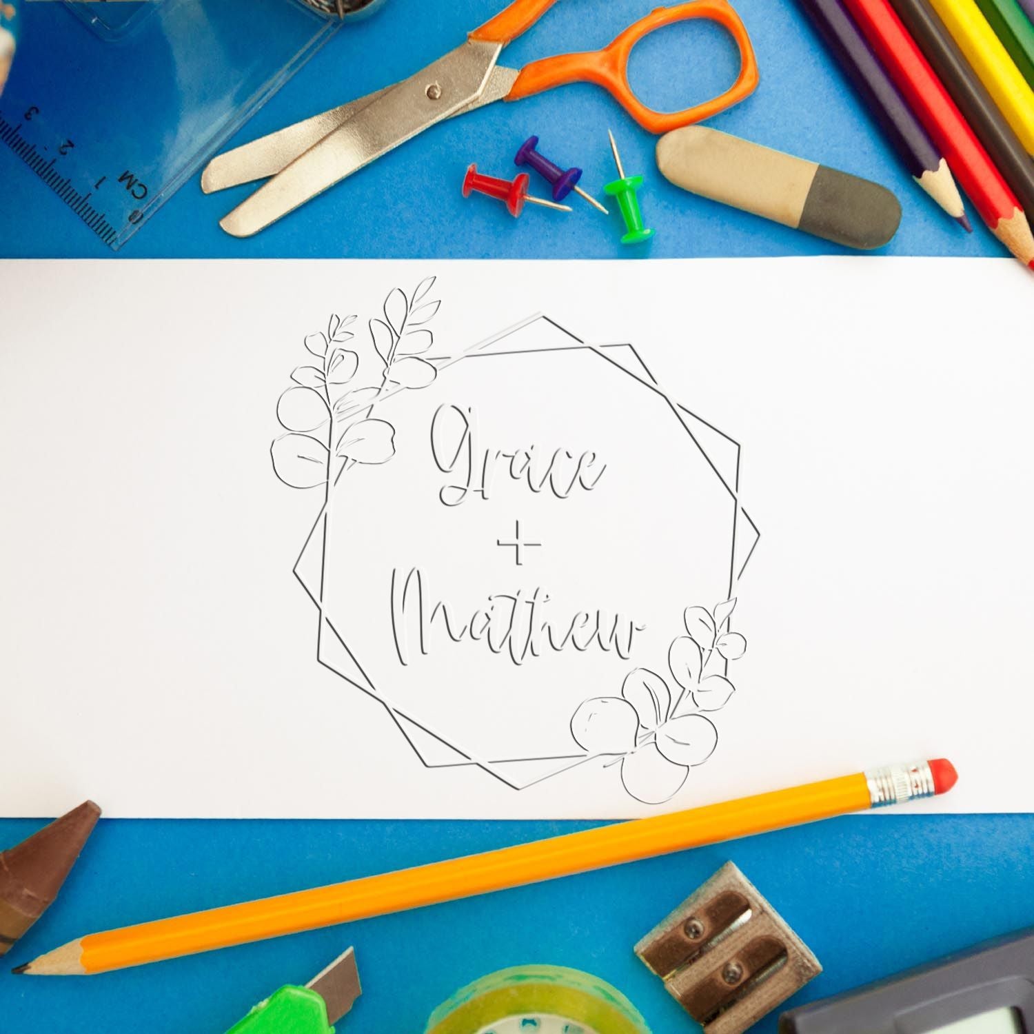 Custom wedding stamp seal reading Grace + Matthew, with floral wreath on white paper, surrounded by blue stationery and pencils.