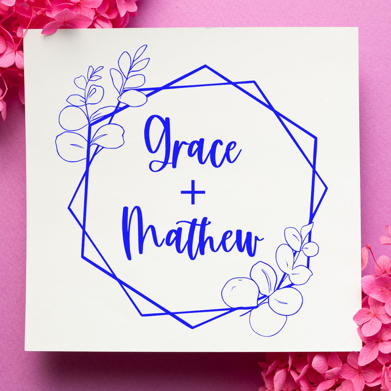 Blue floral custom wedding stamp seal reading Grace + Matthew on a white square, framed by pink flowers.