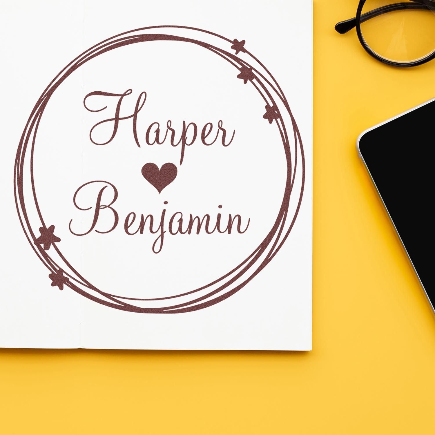 Brown custom rubber stamp seal reading Harper and Benjamin with heart, on white paper against a yellow background with glasses.