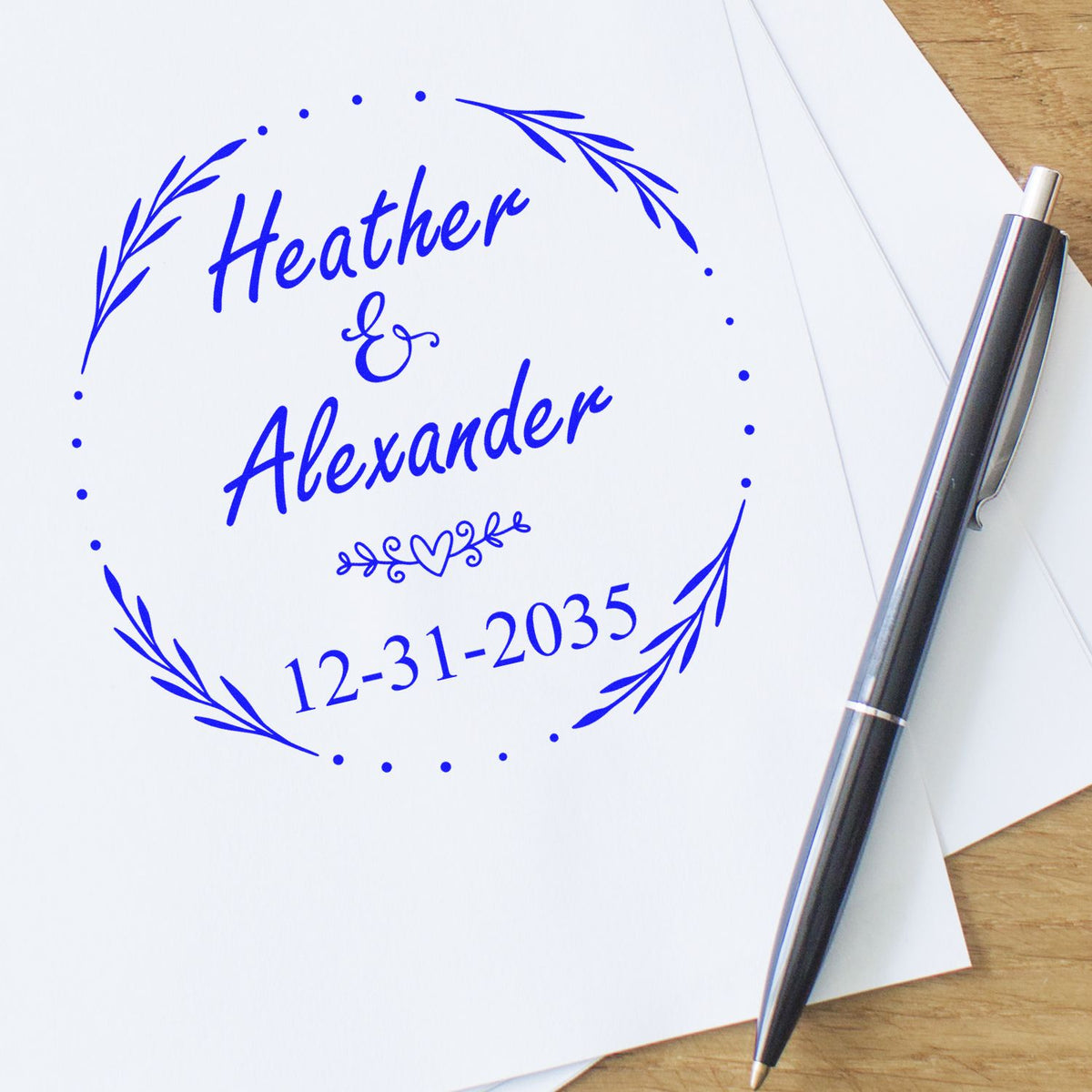 Blue custom wedding stamp seal with Heather & Alexander, laurel wreath, and 12-31-2035 beside a silver pen.