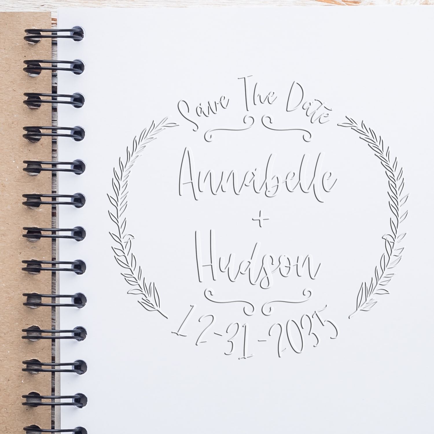 White embossed stamp seal with black wreath, “Save The Date Annabelle + Hudson 12-31-2025,” on a tan spiral notebook.