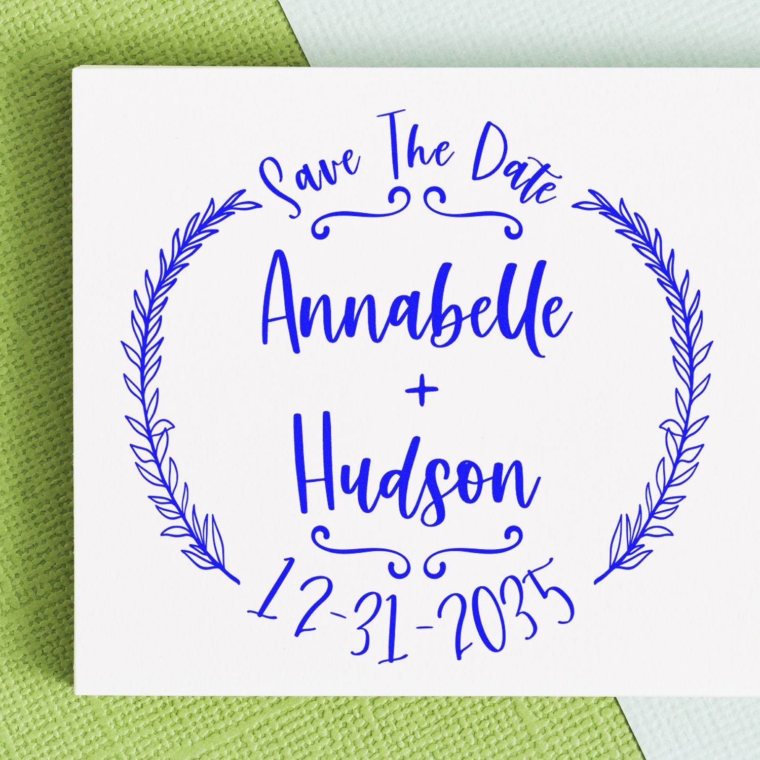 Blue custom save-the-date stamp seal with wreath border for Annabelle and Hudson, dated 12-31-2035, on white card.